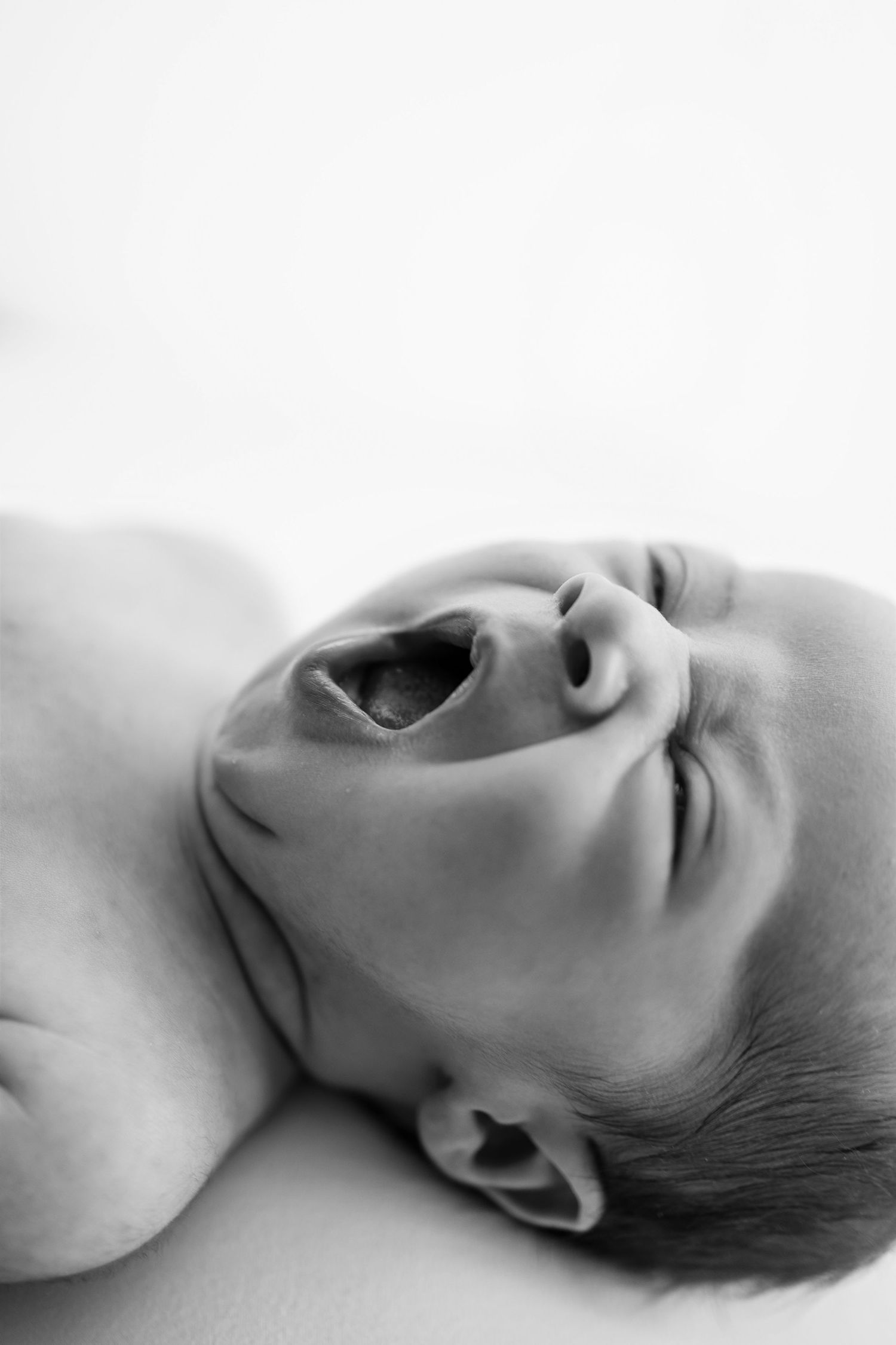 Black and white close-up photo of a newborn baby crying with mouth wide open, lying on a white surface.