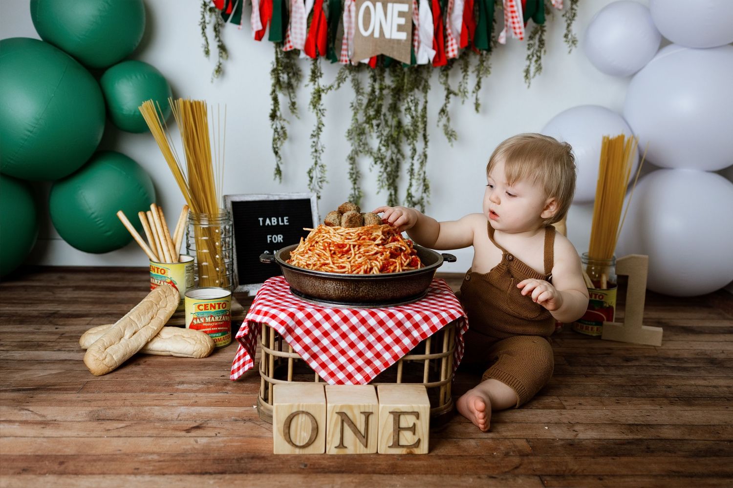 First birthday party setup with green and white balloons, rustic wooden blocks, and red checkered decor on wooden floor.