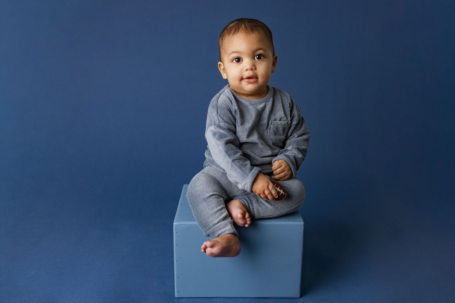 Child in gray sweater sitting on blue cube against navy backdrop in studio portraits.