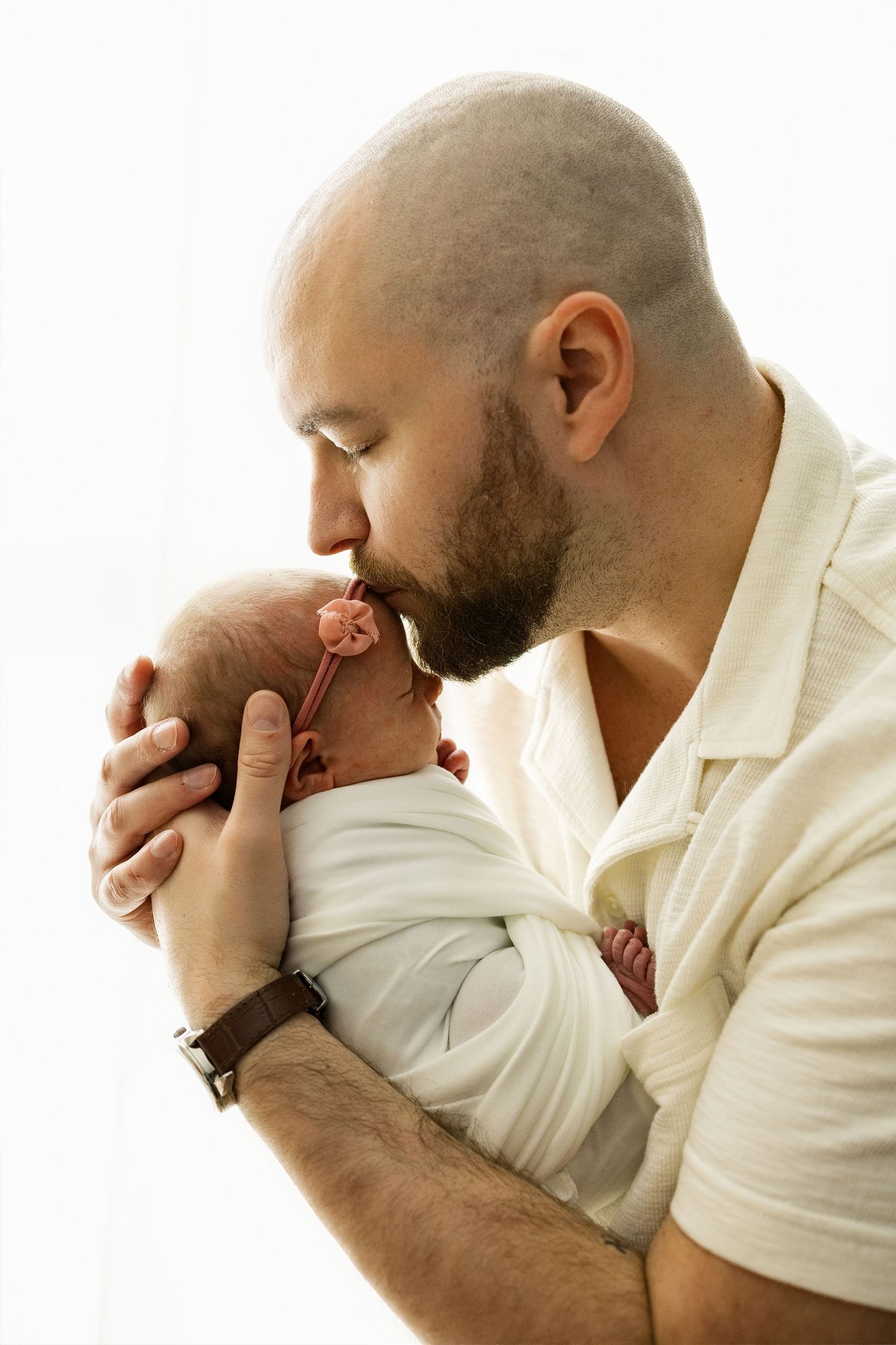 Tender moment captured in soft lighting as parent kisses sleeping newborn wrapped in white.