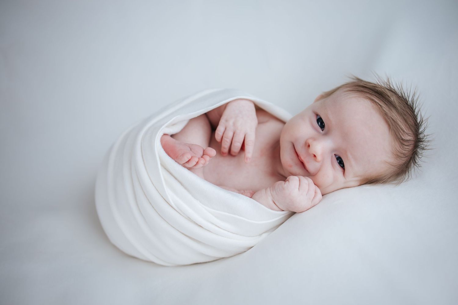 Newborn baby wrapped in soft white blanket laying on light background.