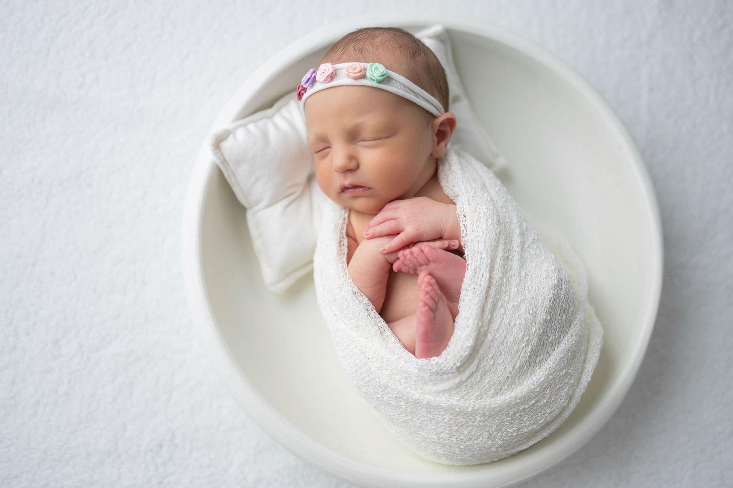 A sleeping newborn baby wrapped in white swaddling cloth lies curled in a white round bowl wearing a floral headband.