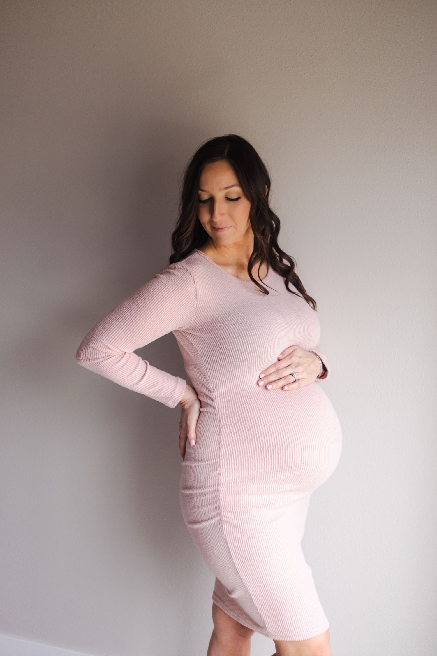 Maternity portrait of woman in pink fitted dress against neutral wall.