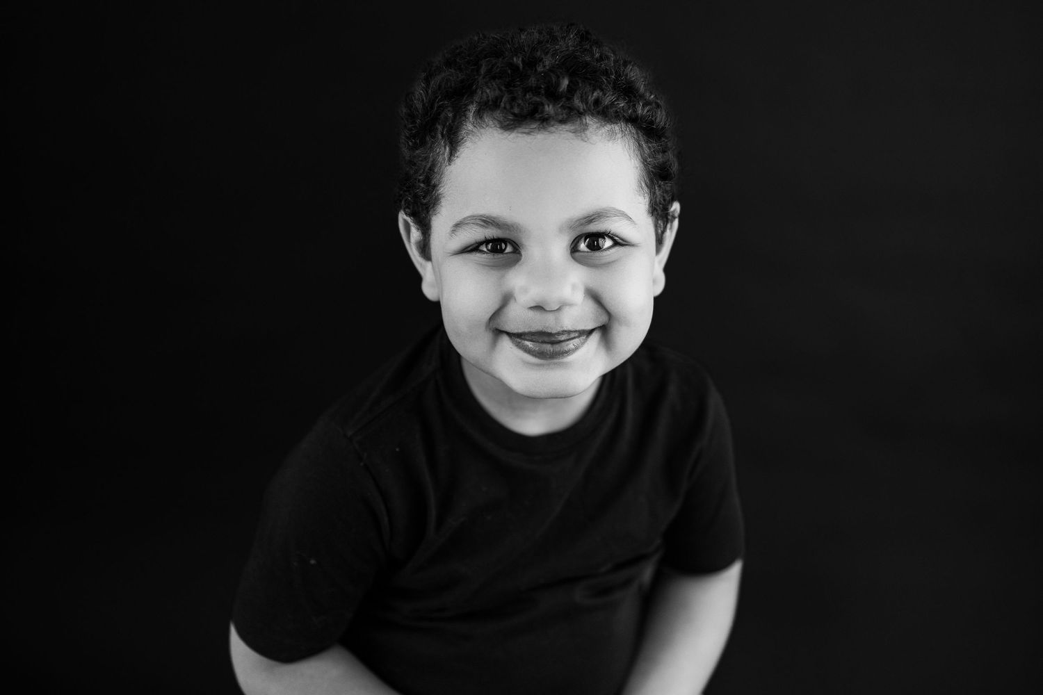 Black and white portrait showing a smiling youth in dark clothing against a dark background.
