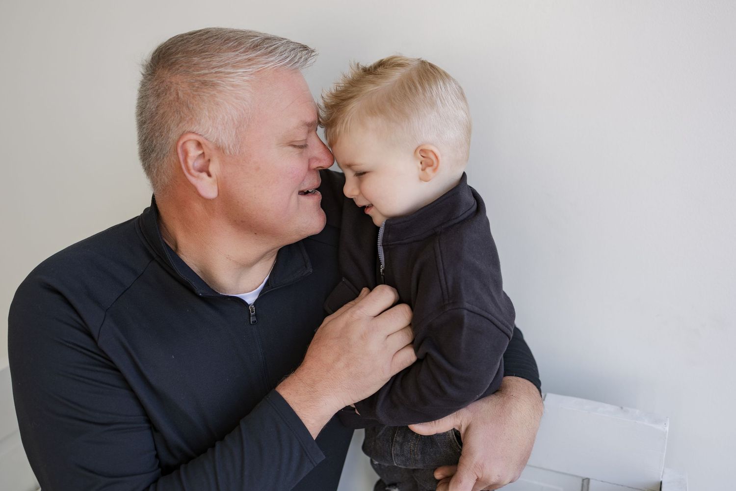 A grandfather lovingly holds his young toddler grandson, touching foreheads in a tender moment, both wearing dark jackets.