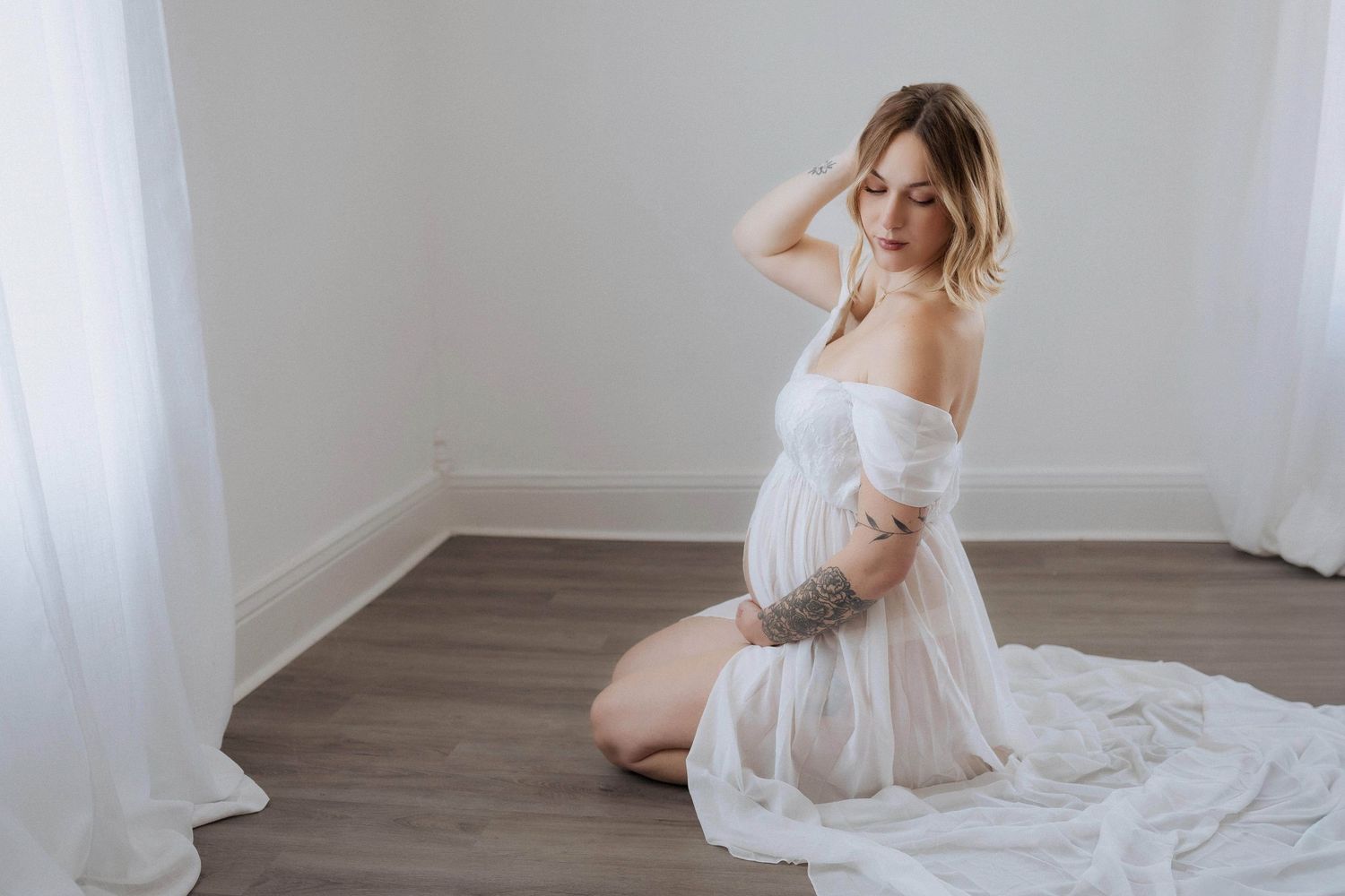 A figure in a flowing white dress sits gracefully on a wooden floor in an ethereal bridal pose.