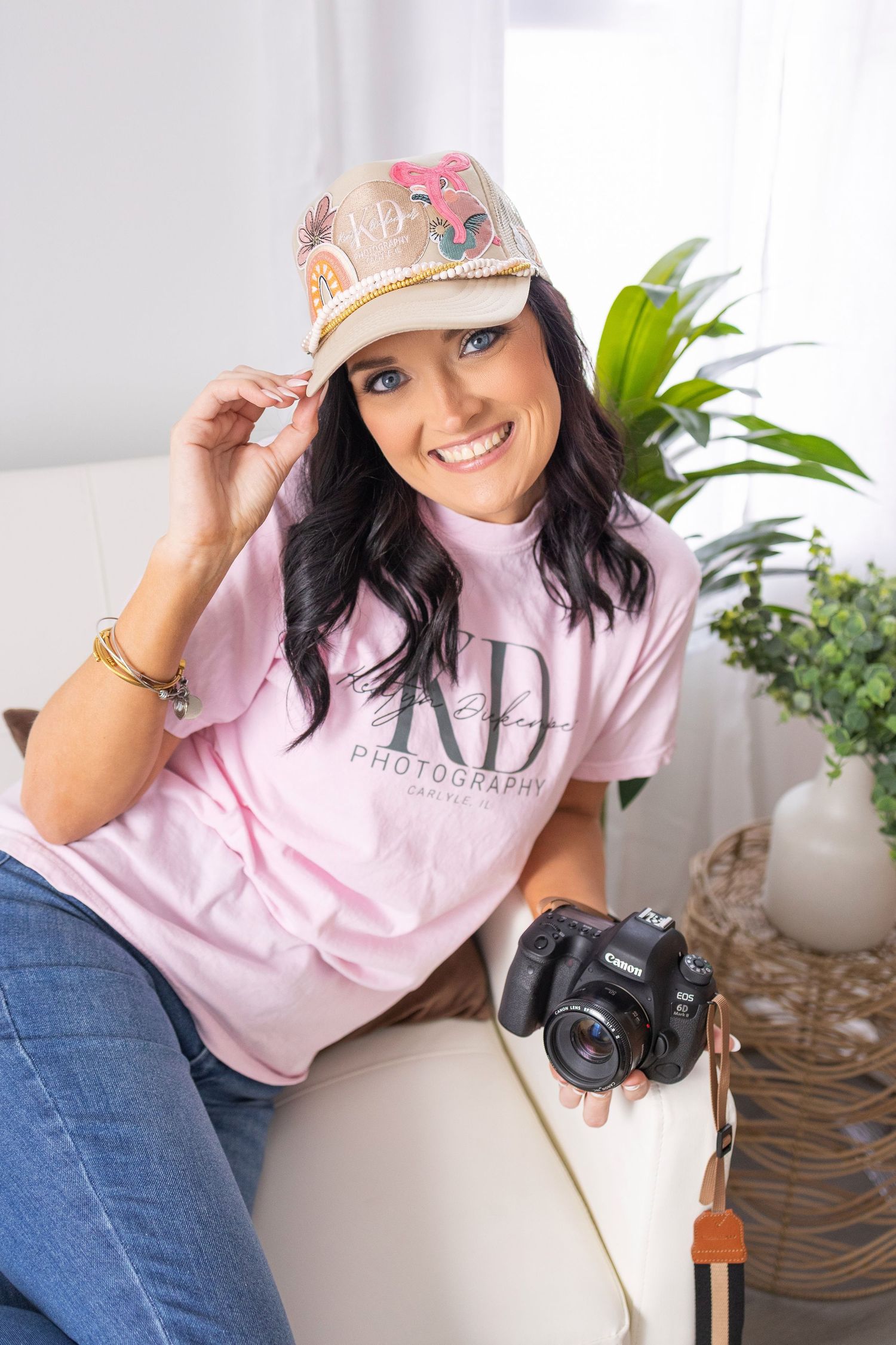Photographer wearing pink t-shirt and decorative cap poses with camera near indoor plants.