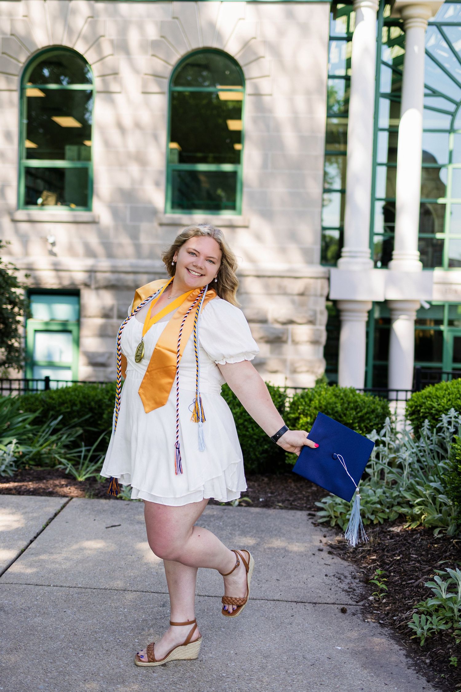 A graduate in a white dress with yellow sash poses happily with diploma outside a building with arched windows.