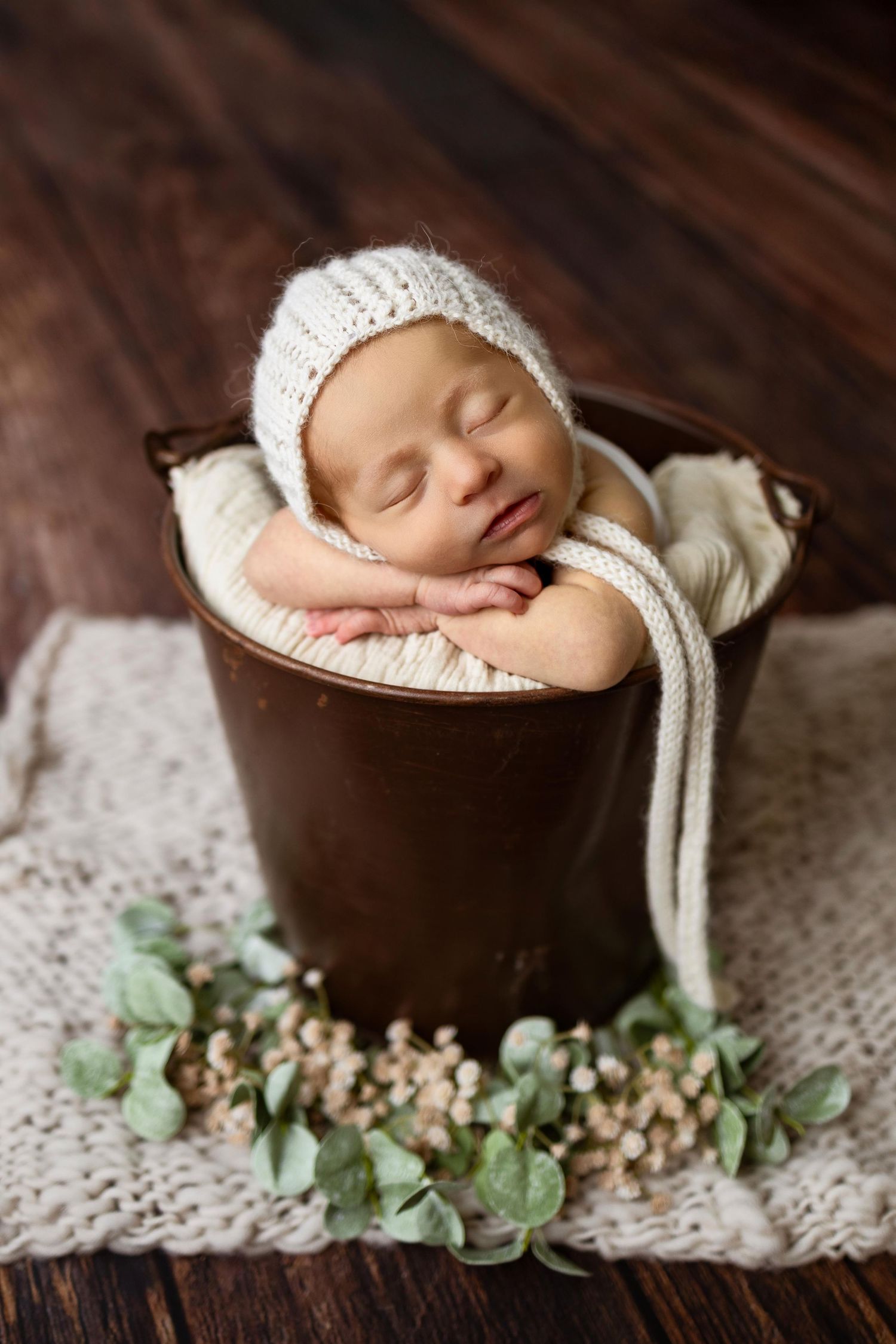 Sleeping newborn in white knit bonnet rests on wooden bowl with eucalyptus leaves and lace blanket in studio setting.