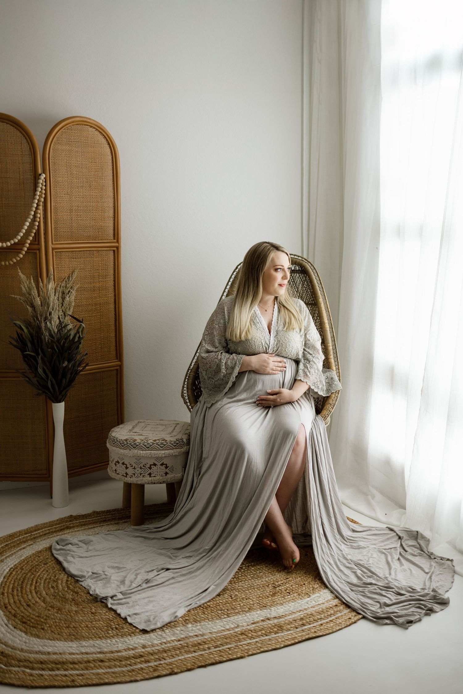 Maternity photoshoot featuring flowing gray dress on jute rug with rattan screen and natural light from window.