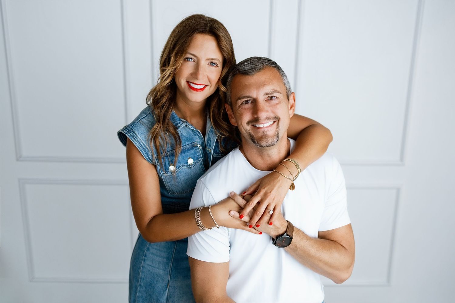 Happy couple posing together, woman in denim dress hugging smiling man in white t-shirt against light background.