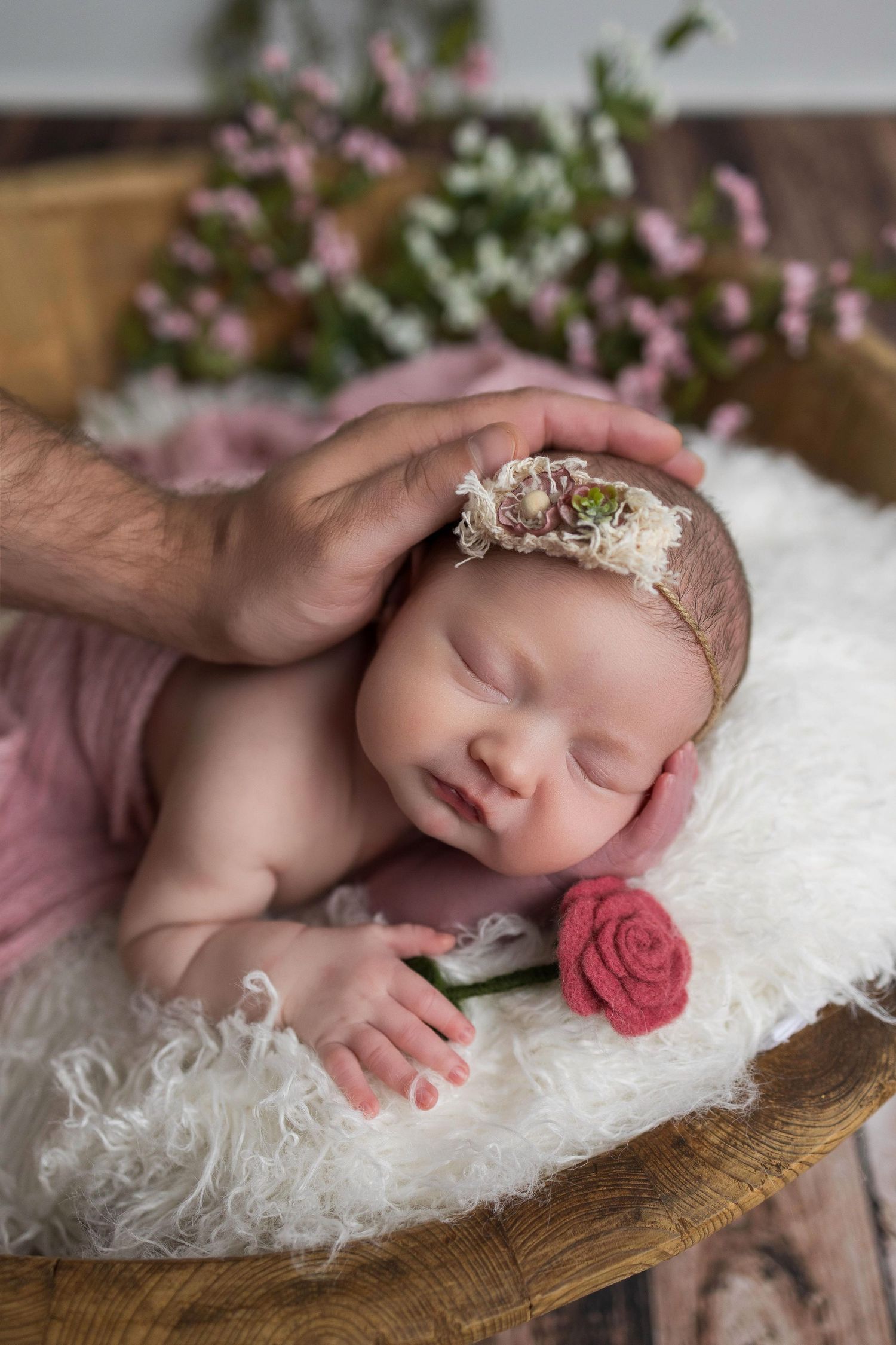 Sleeping newborn on white fur blanket with rose pink headband and floral decorations in wooden basket.