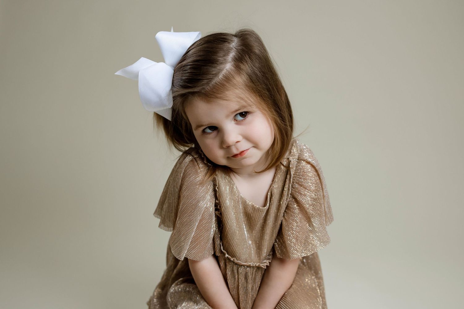A child wearing a brown dress poses against a neutral background in a natural portrait style.