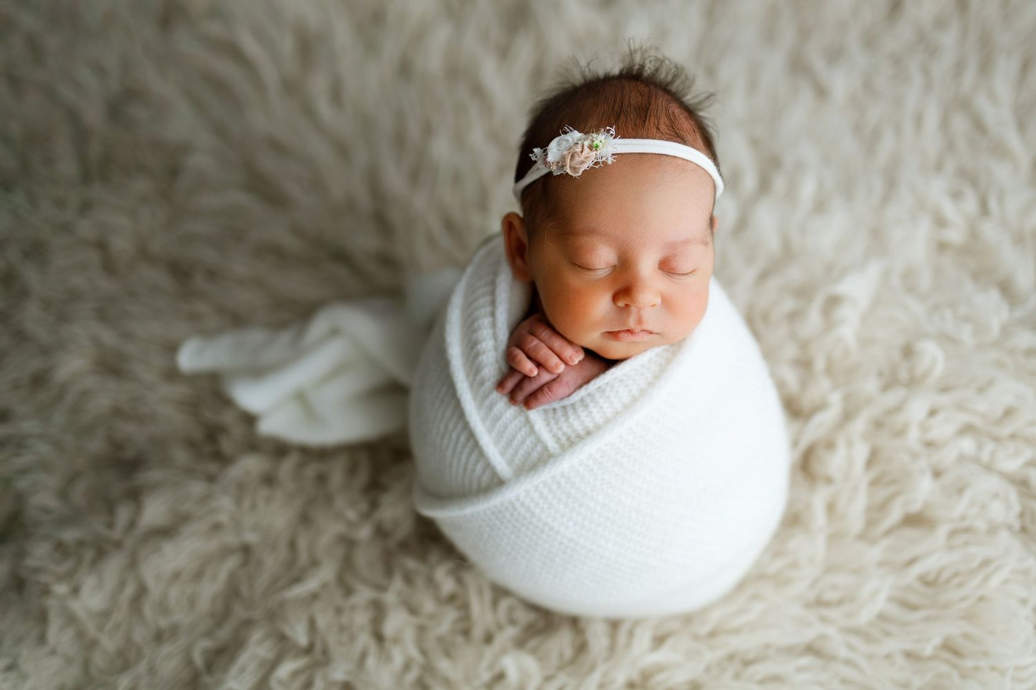 Newborn baby girl swaddled in white wrap with floral headband, sleeping peacefully on fluffy cream fur rug.