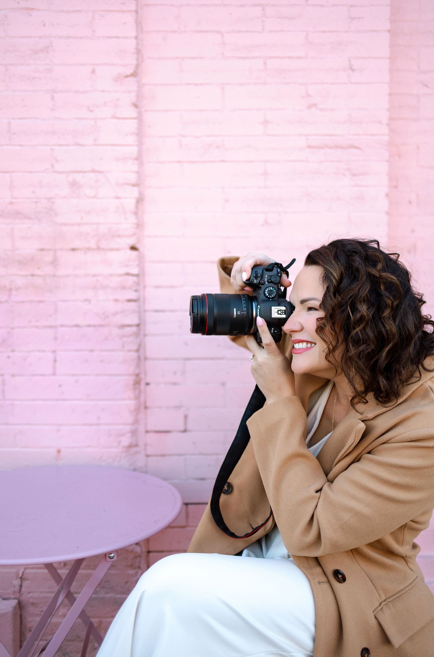 Professional photographer in tan coat takes photos with DSLR camera against pink brick wall.