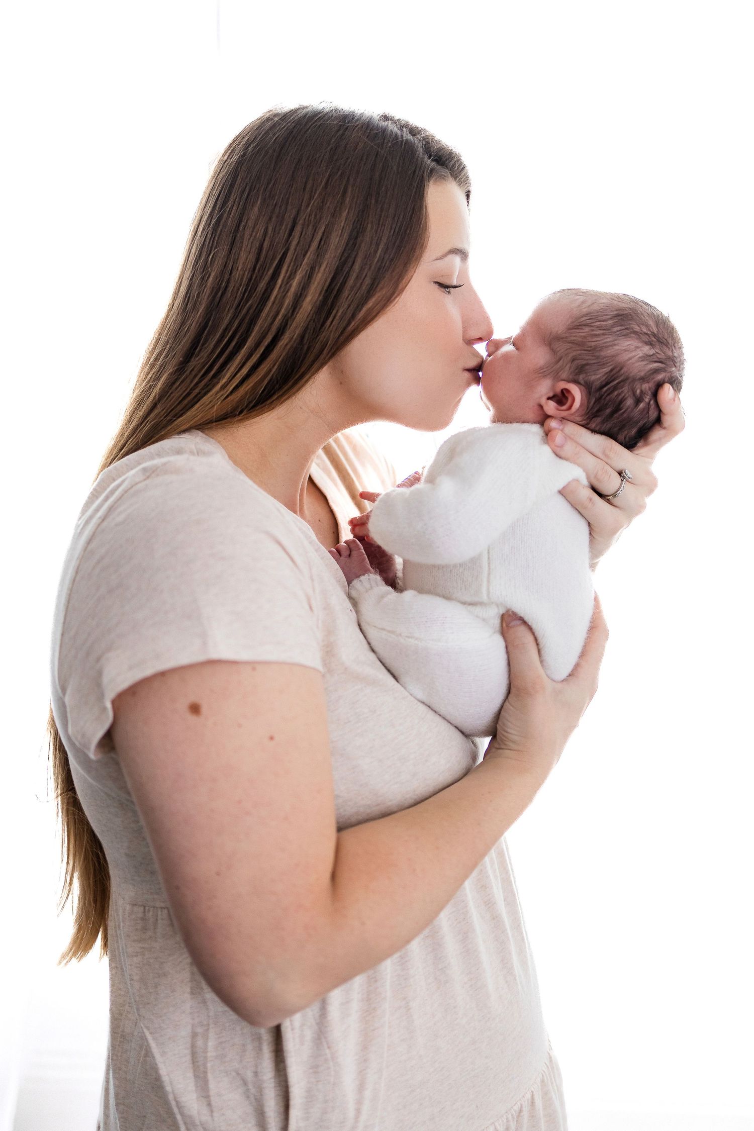 A tender moment shows a woman in a beige shirt gently kissing a newborn baby wrapped in white.