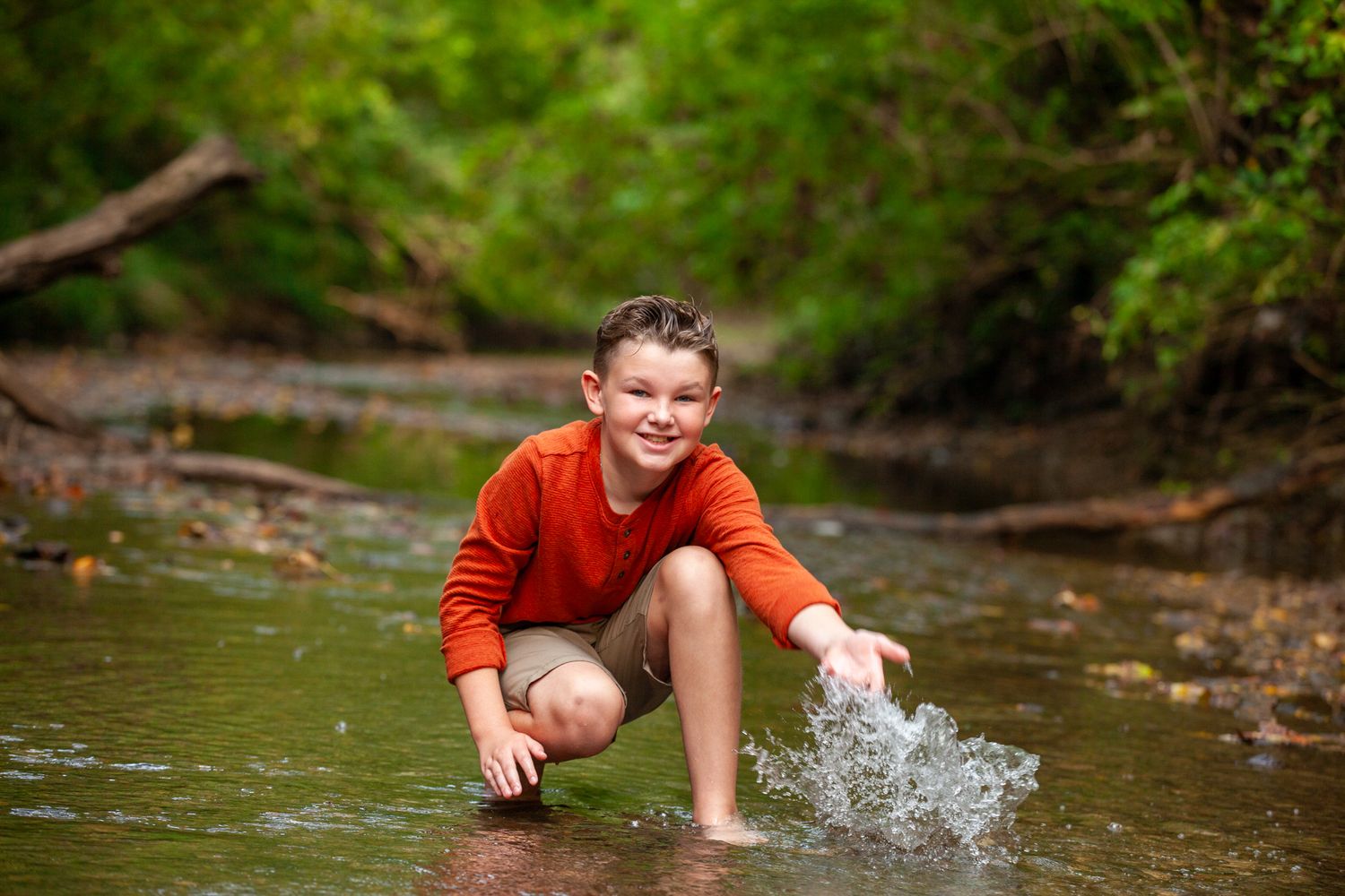 A child in an orange shirt plays and splashes in a shallow creek on a summer day.