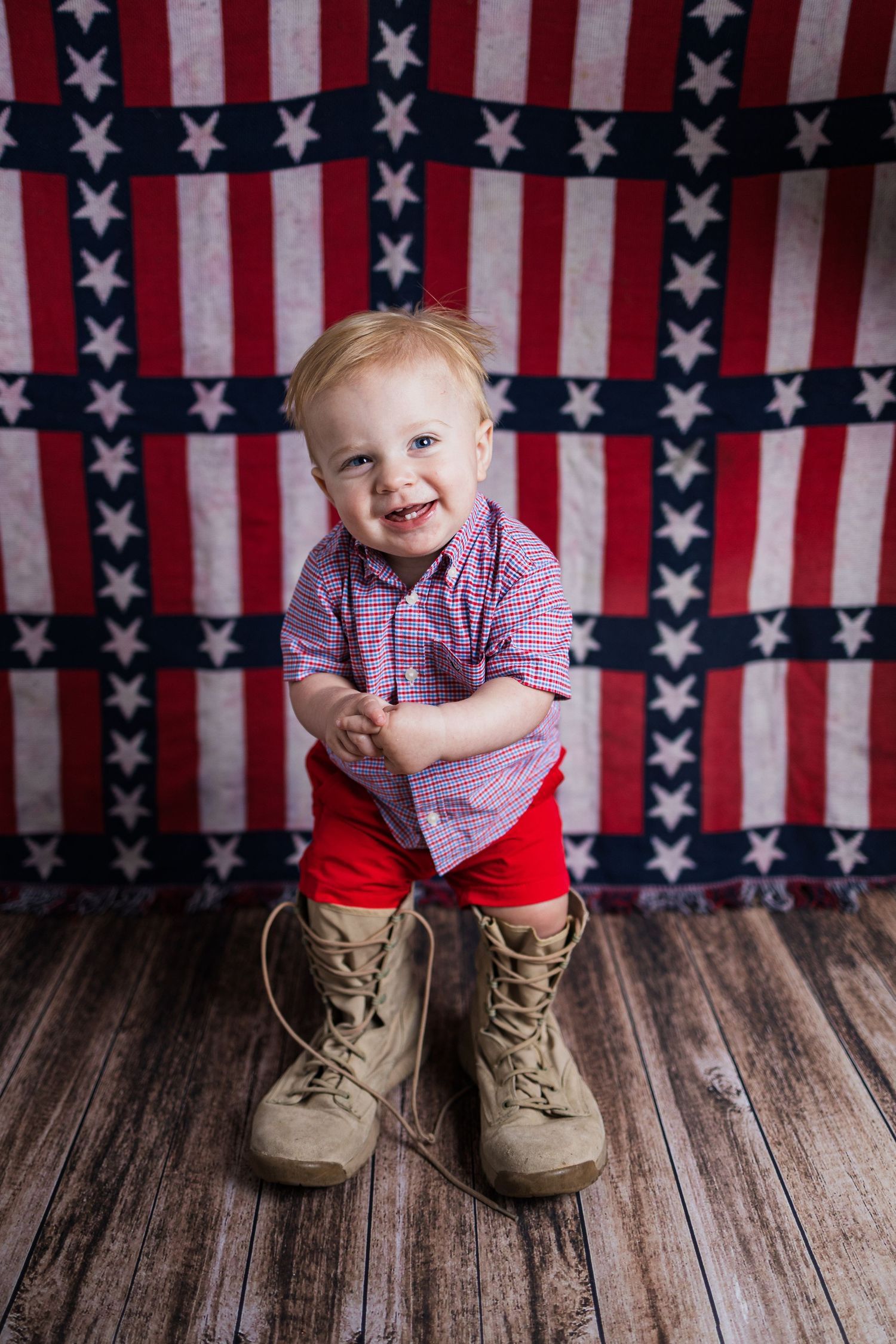 Small child wearing red shorts and checkered shirt stands against American flag backdrop on wooden floor.