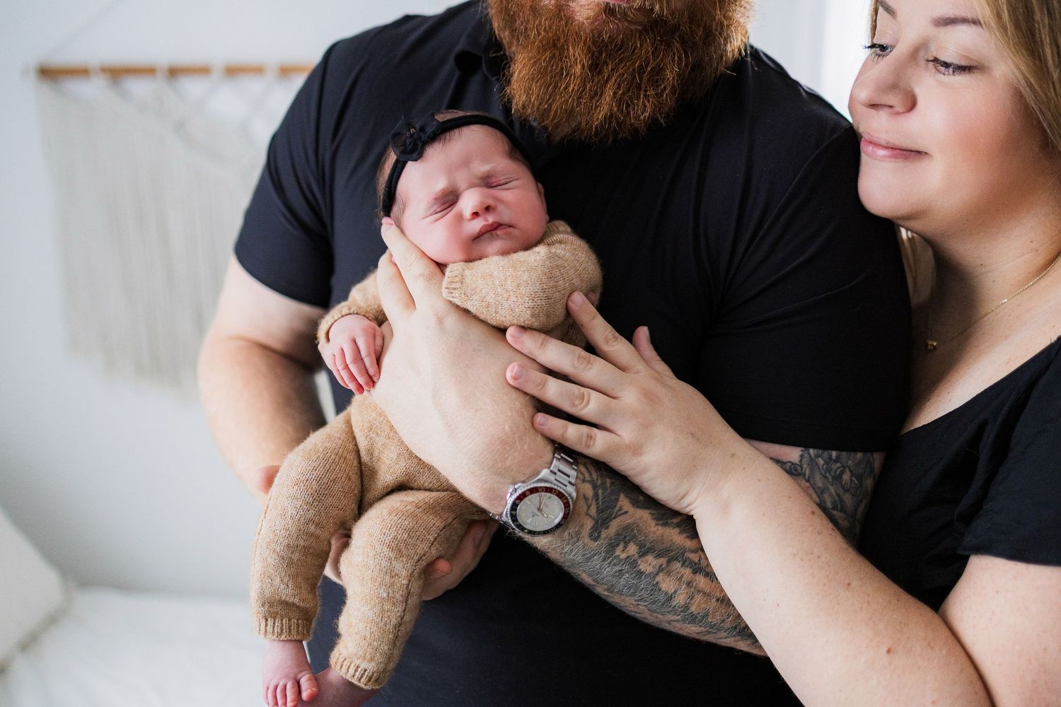 Parents hold their newborn baby wrapped in tan blanket while sitting on white sofa.