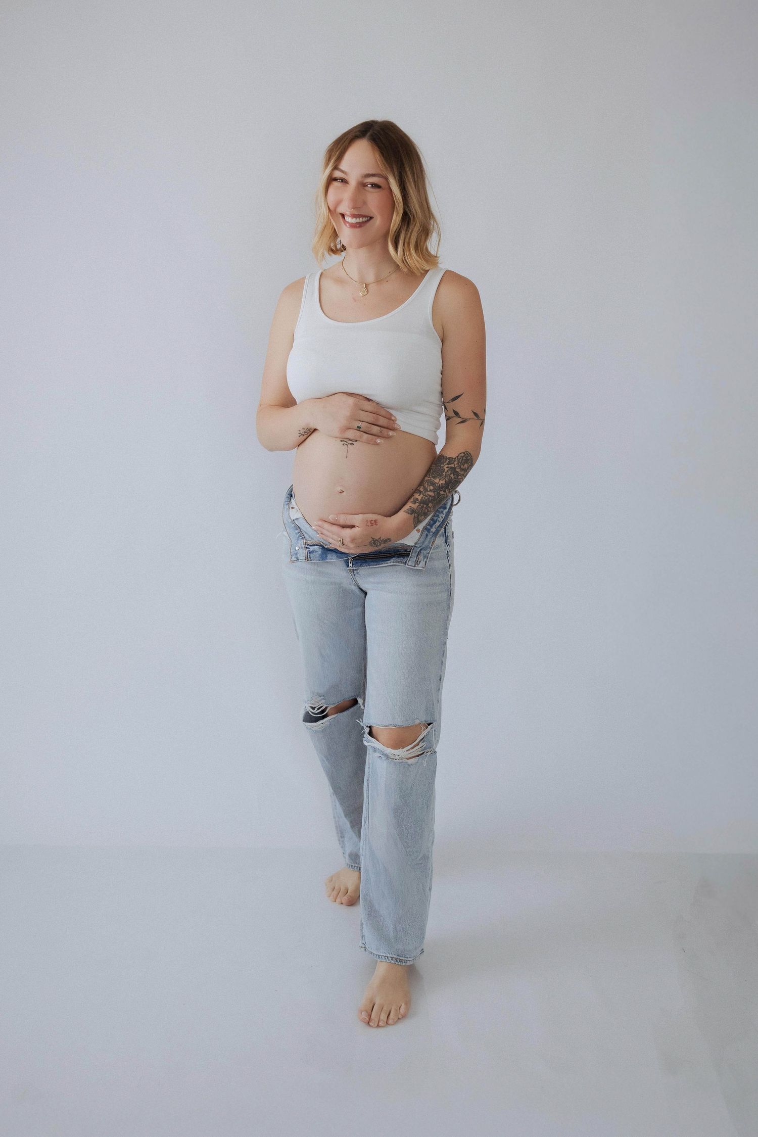 A pregnant person wearing a white tank top and ripped jeans poses barefoot against a light gray backdrop.