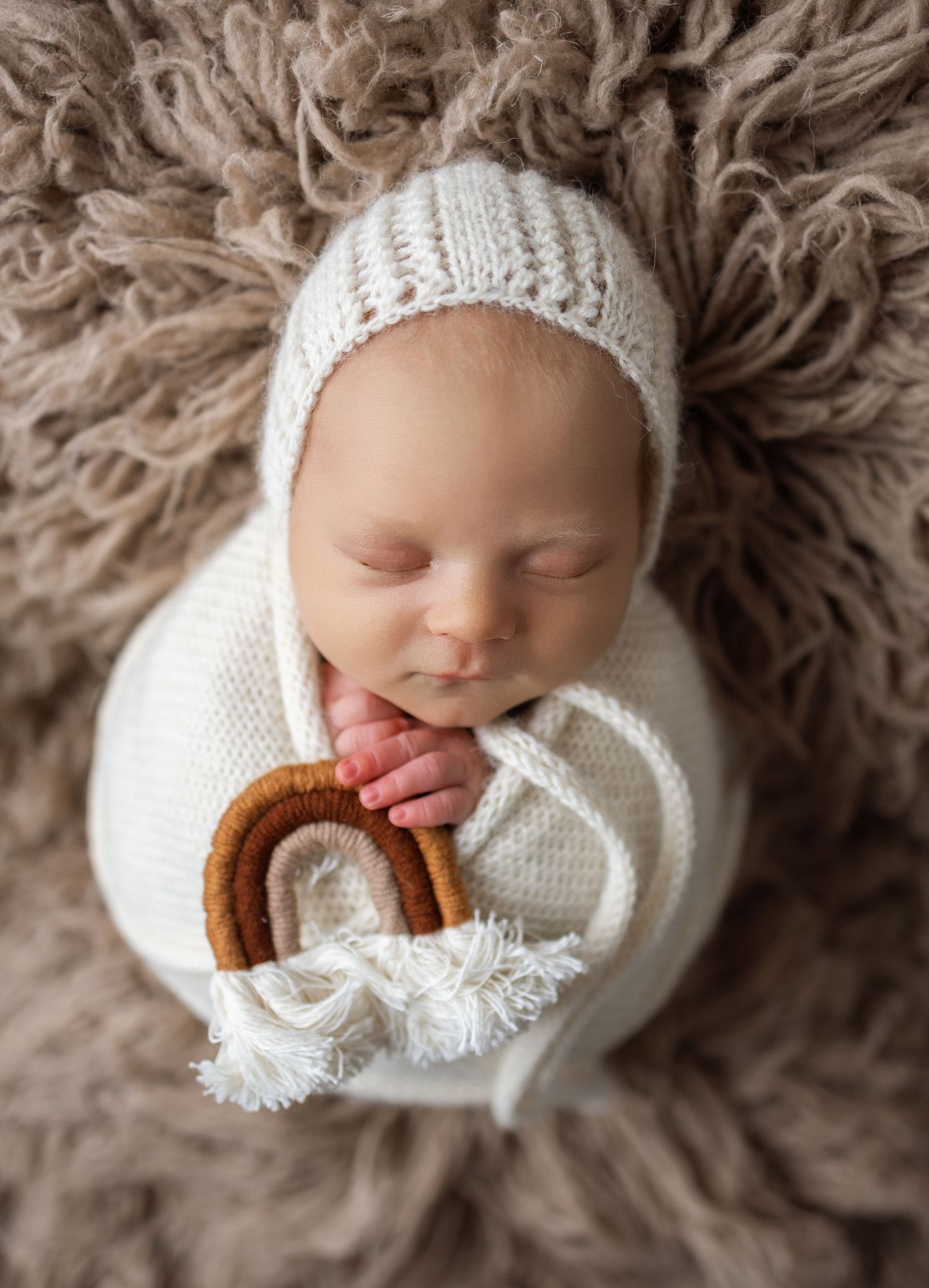 A sleeping newborn wrapped in white knit blanket lies on soft brown fur with a small pretzel in their hand.