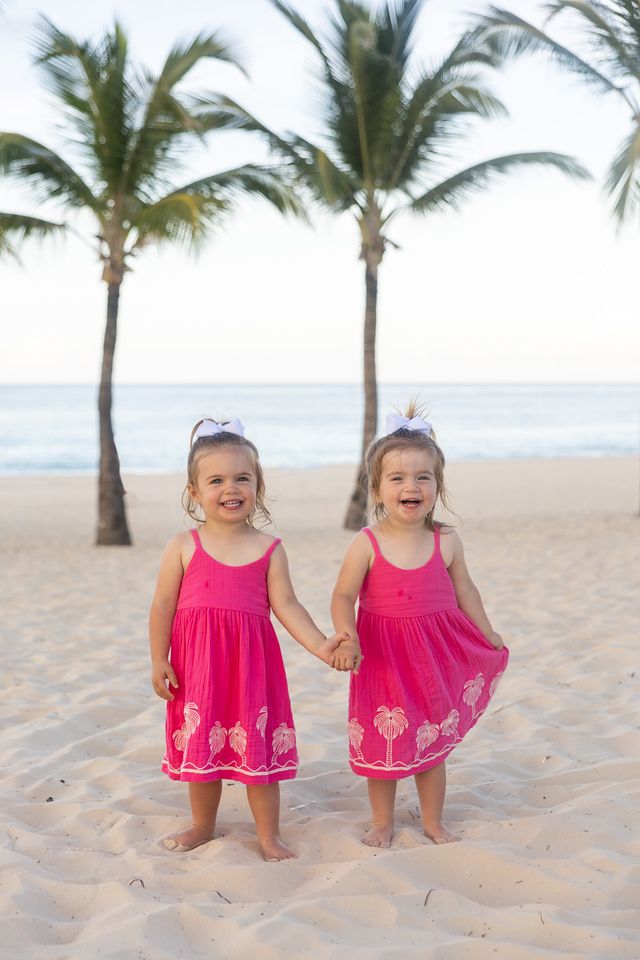 Two young children in matching pink dresses hold hands while standing on a tropical beach with palm trees.
