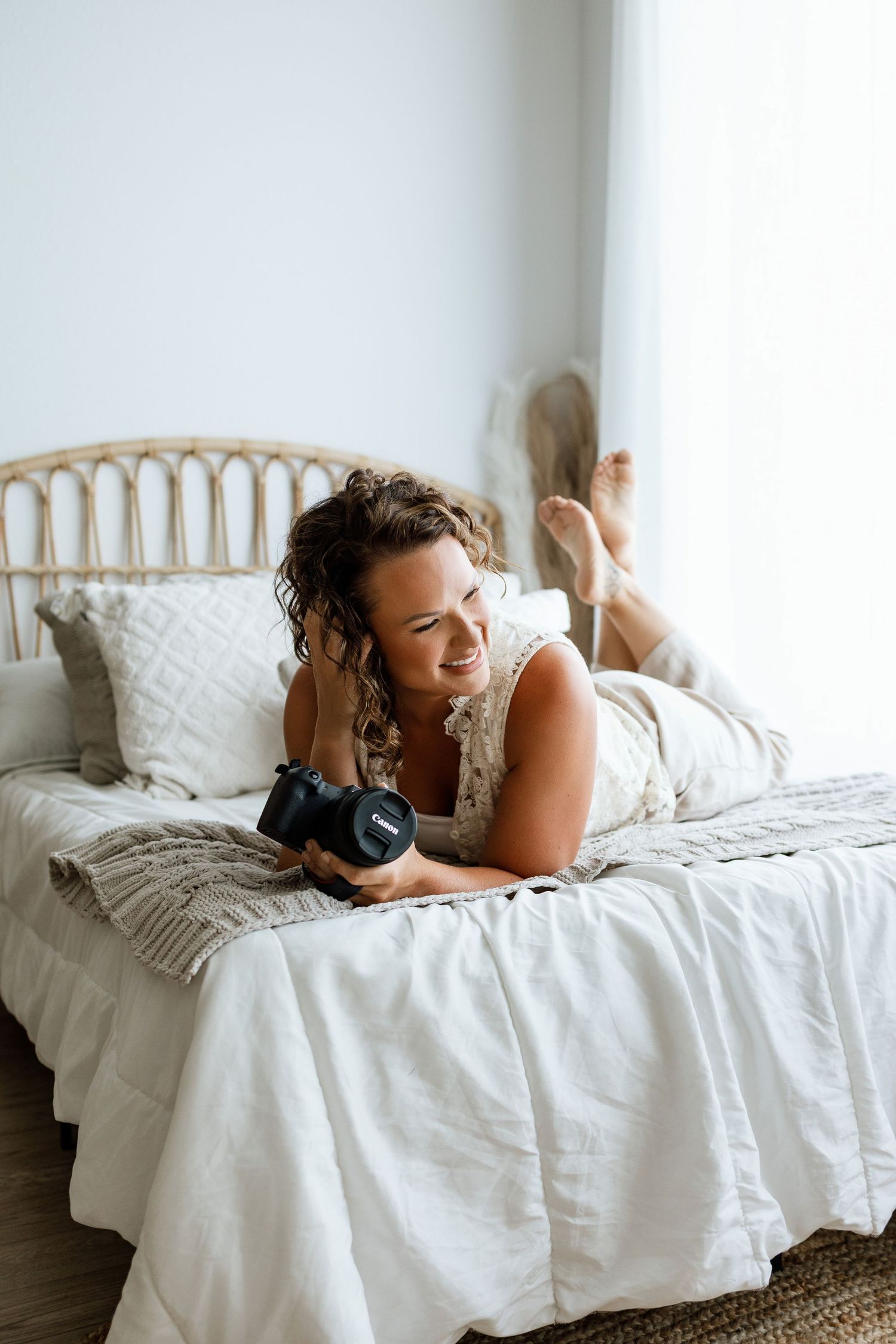 Woman lying on white bed holding a camera, smiling in a bright bedroom with rattan headboard.