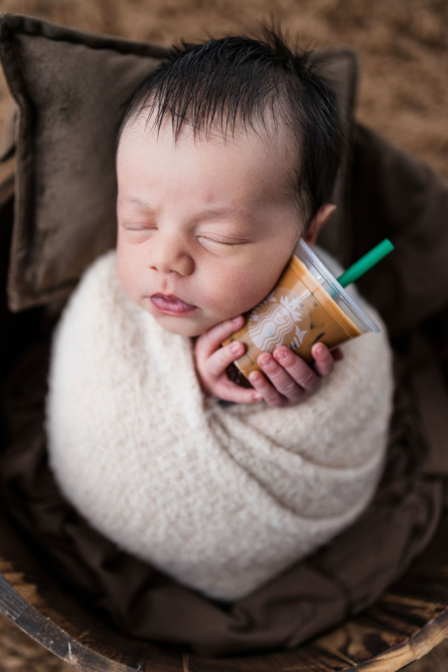 Sleeping baby wrapped in white blanket holding Starbucks cup in unique newborn photography session.