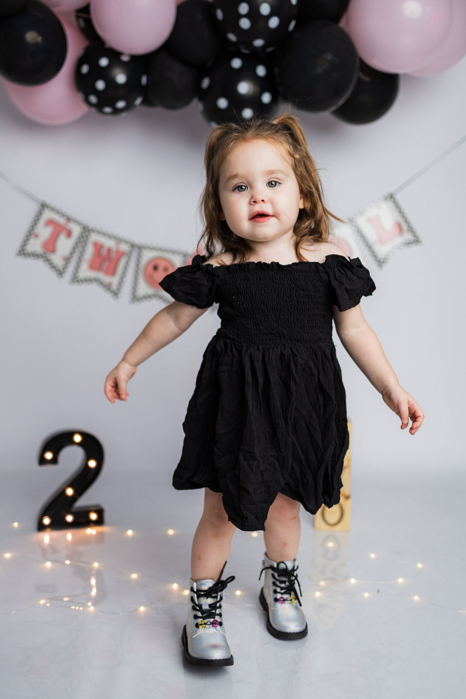 A small child in a black ruffled dress and silver shoes poses against a backdrop with pink and black balloons.
