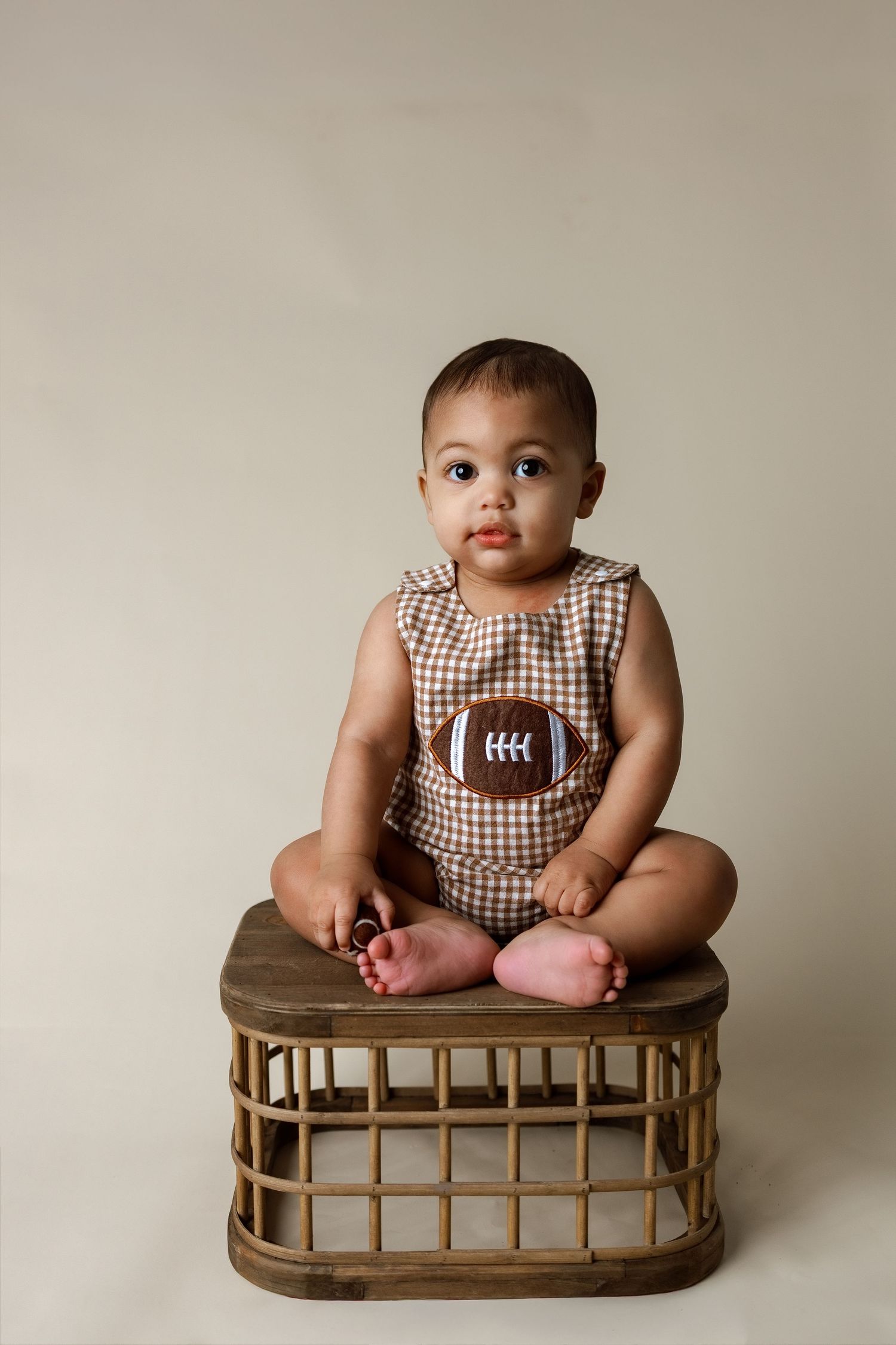 A baby in a brown gingham romper sits on a wooden bench prop during a professional photo session.