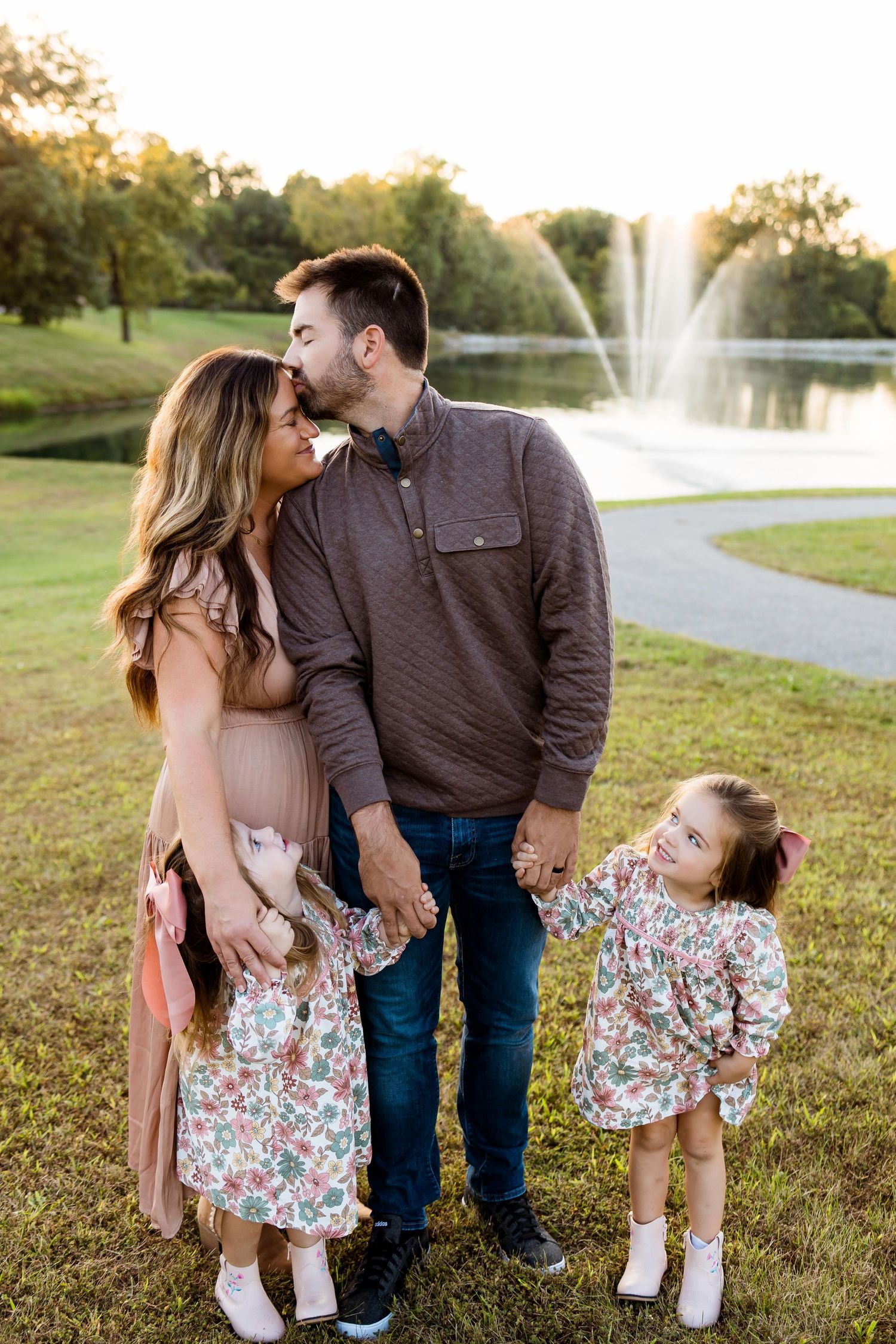 A family shares tender moments together on grassy lawn near fountain during golden hour sunset.