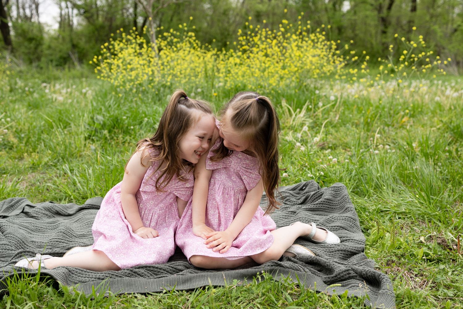 Two children in matching pink dresses share sweet moment on blanket outdoors.