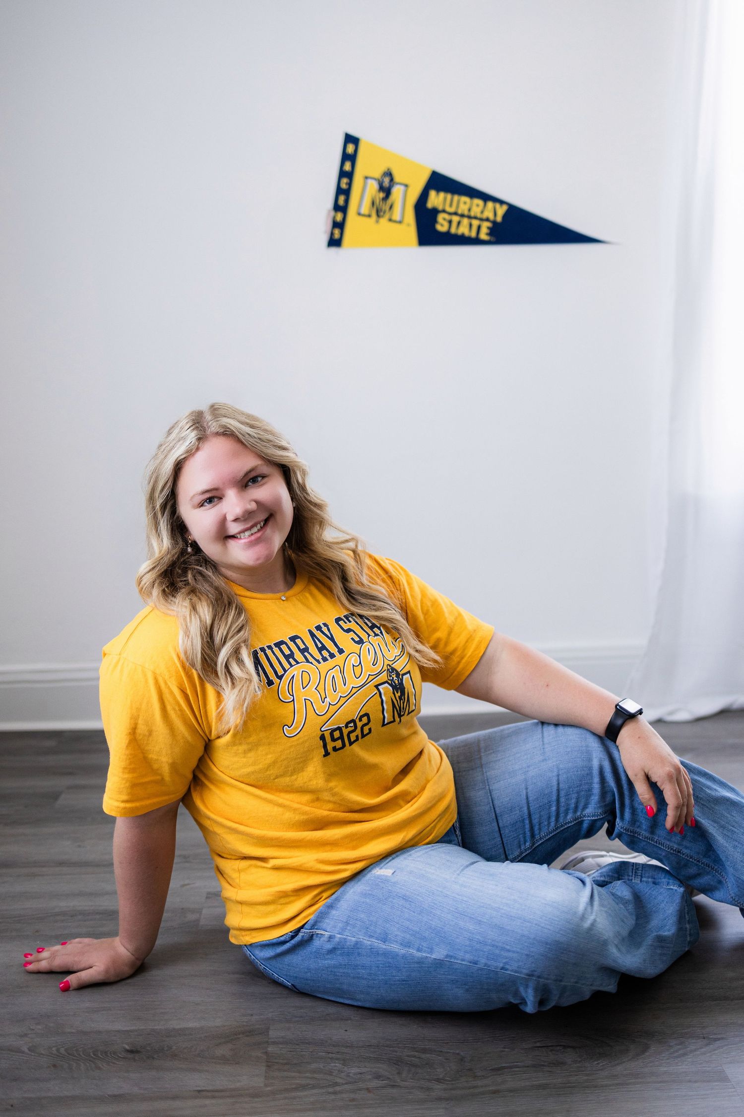 A college student in yellow Murray State shirt sits casually on hardwood floor with university pennant on wall.