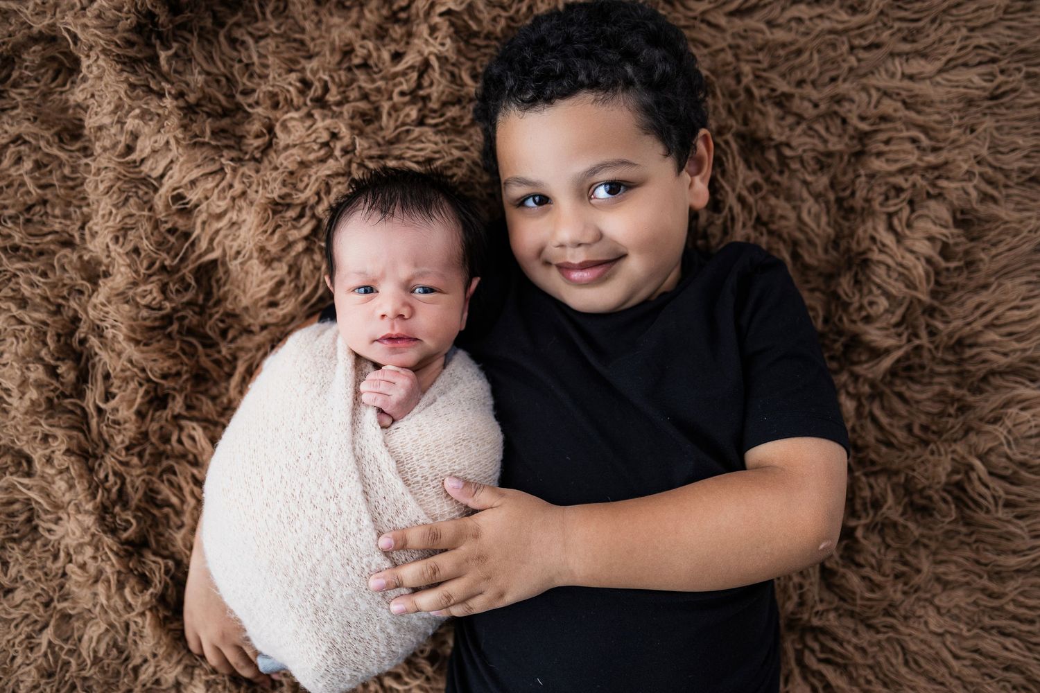 Two children sitting together on a brown fur blanket with one wrapped in white swaddling.