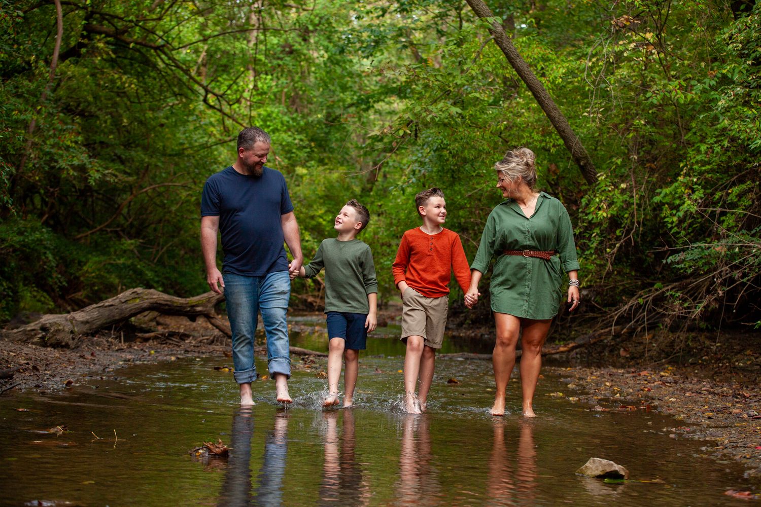 A family walks along a wooded creek bed with large fallen trees and rocks while exploring nature together.