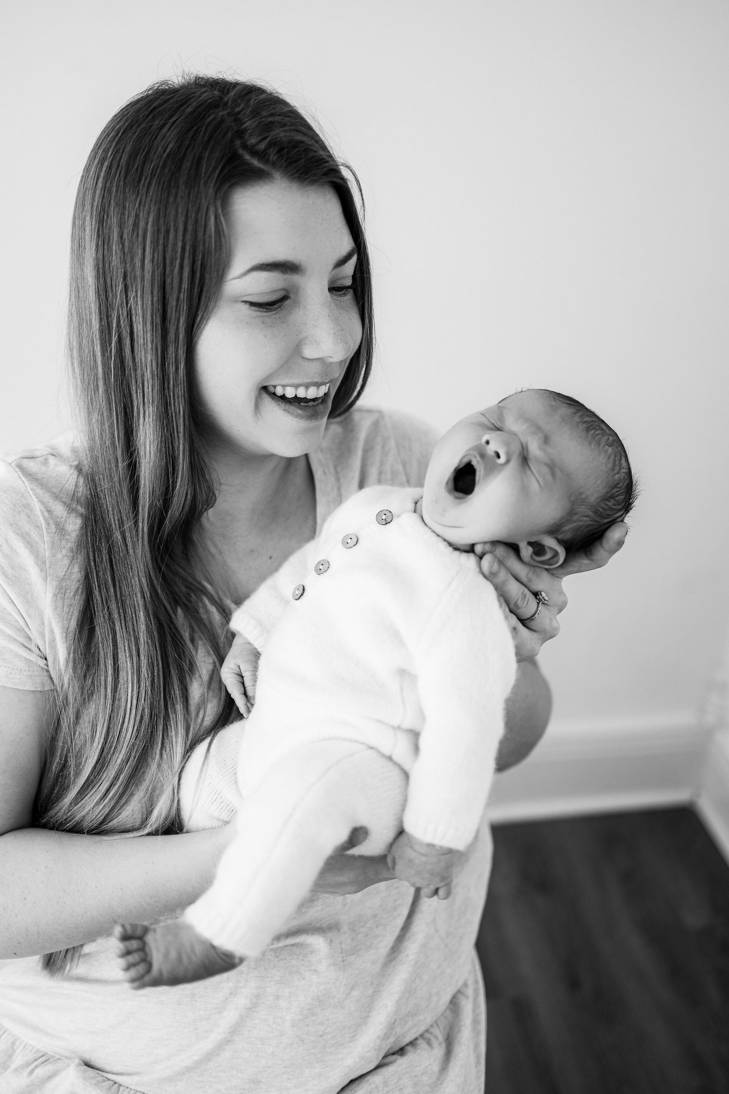 A joyful black and white portrait of a mother holding her yawning baby in a white onesie.