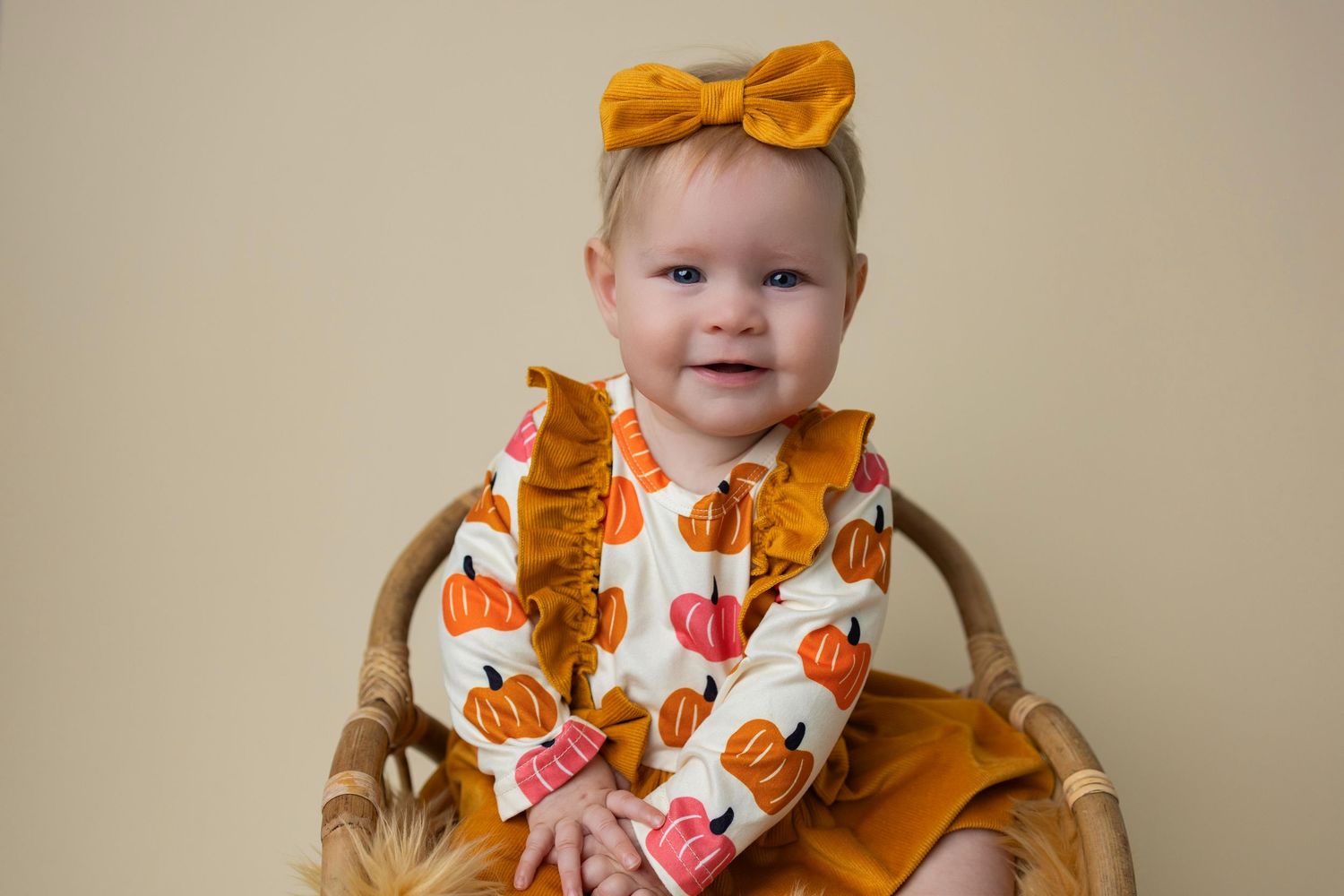 Baby wearing mustard yellow bow and floral outfit sitting in wooden chair against neutral background.