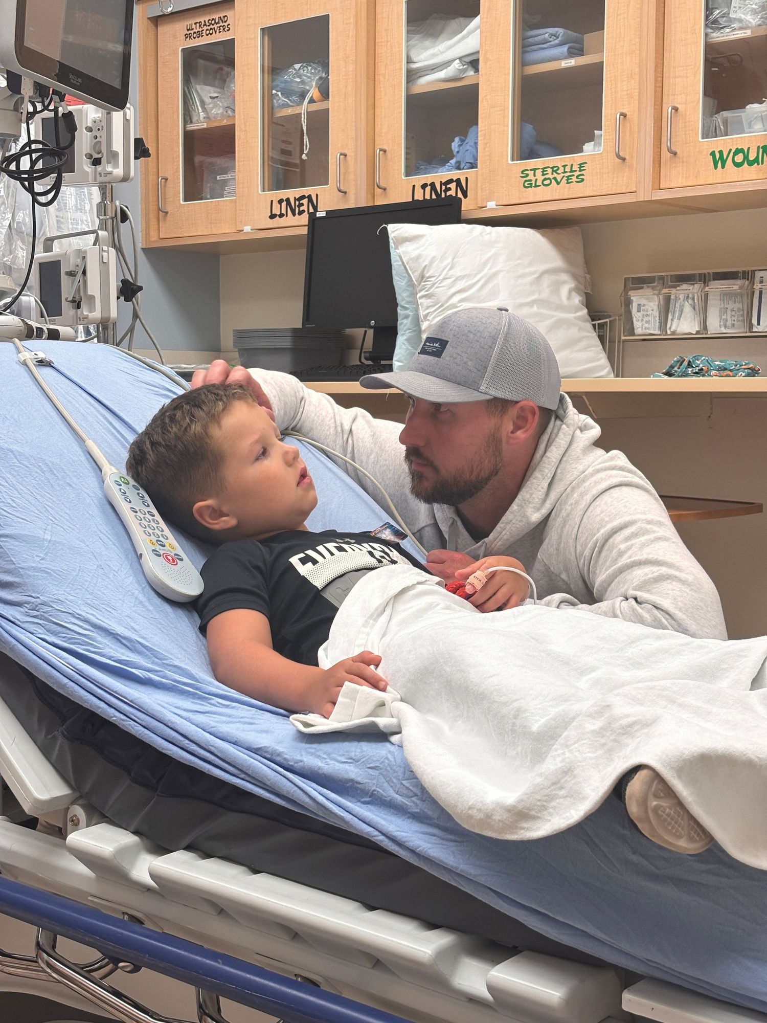 A man in a white hoodie tenderly comforts a young child lying in a hospital bed in a medical room.