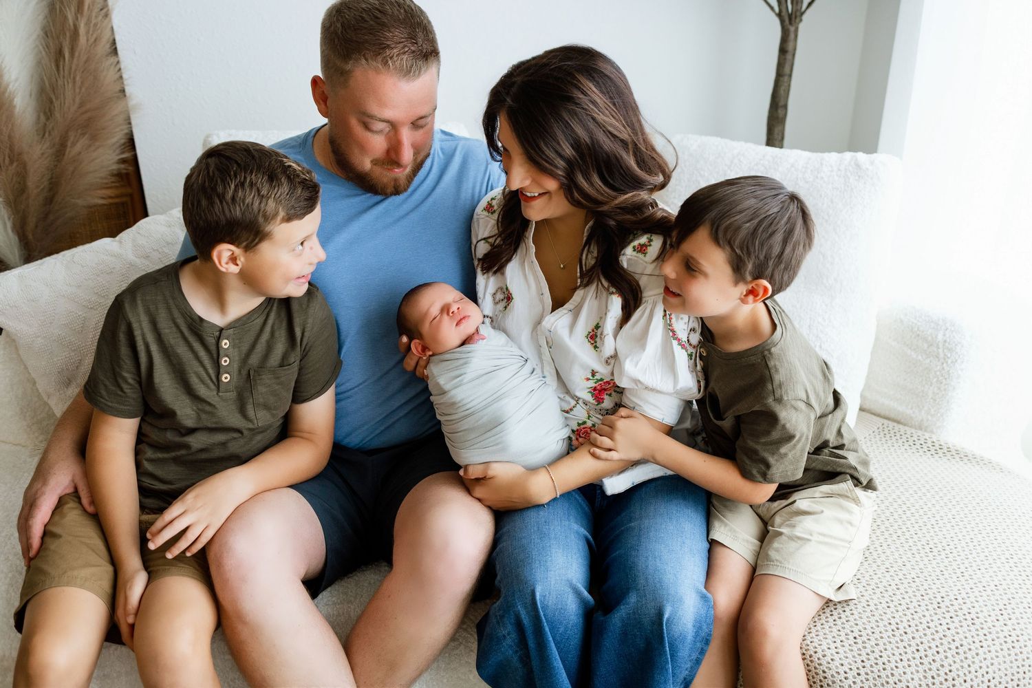 Family of five sitting on couch, parents holding newborn baby while two young boys look on lovingly.