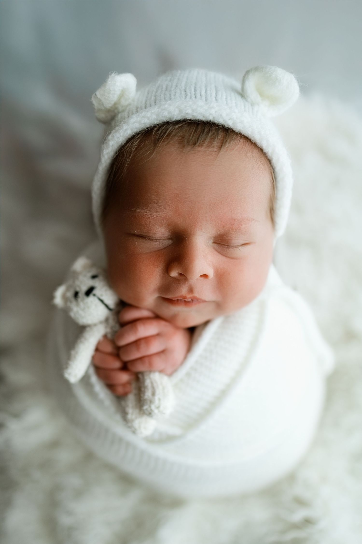 Sleeping newborn baby in white bear bonnet hat holding a tiny knitted teddy bear, wrapped in white.