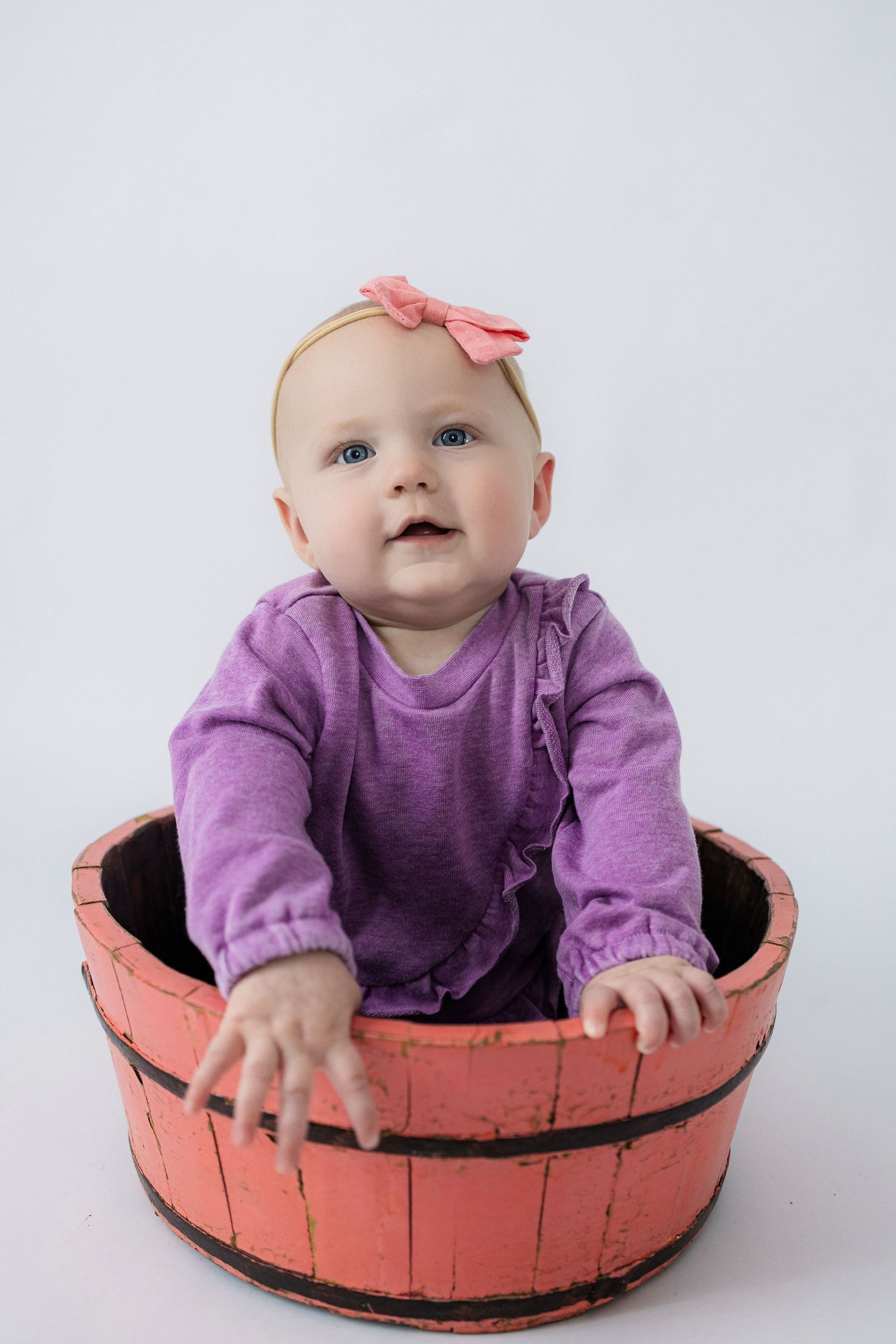 Smiling baby wearing purple sweater sitting in vintage coral-colored wooden barrel.
