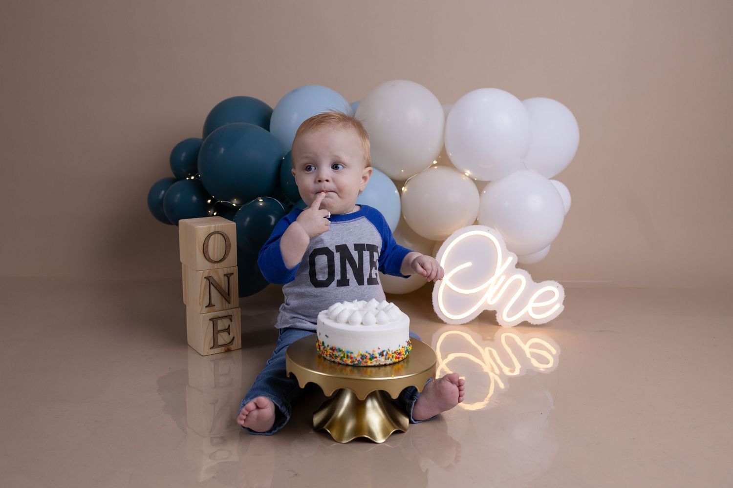 First birthday celebration setup with blue and white balloons, cake smash scene, and neon 'One' sign on beige backdrop.