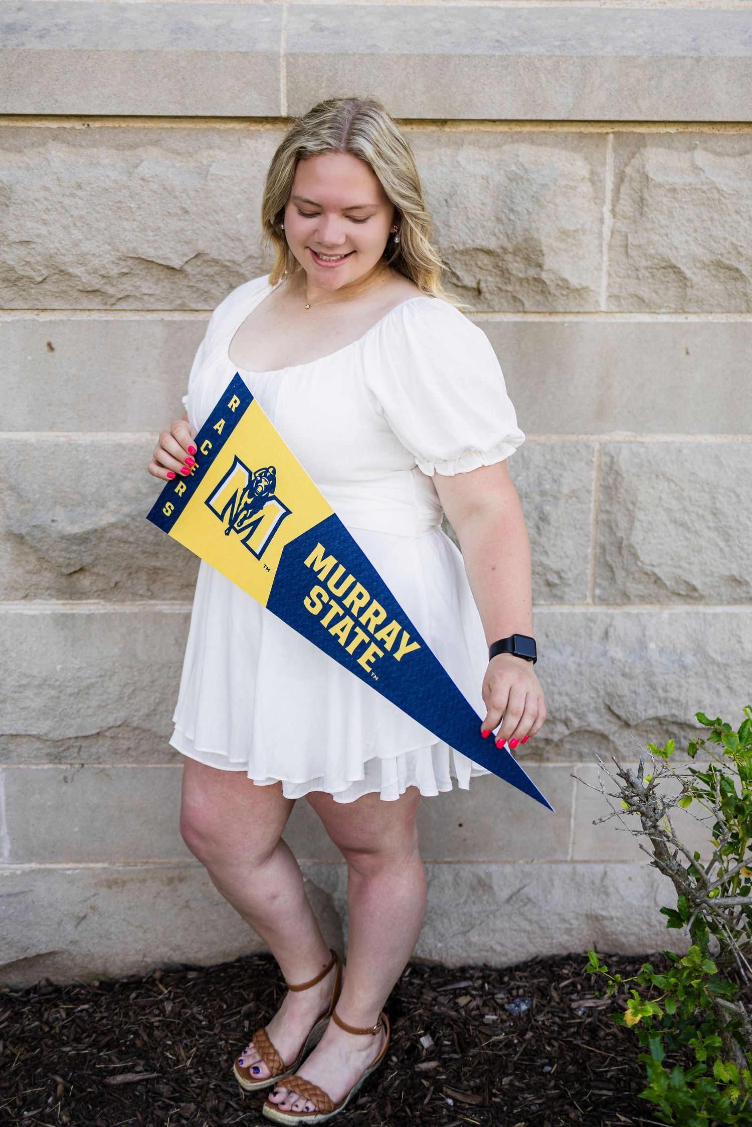 Graduate in white dress holding Murray State university pennant poses against stone wall.