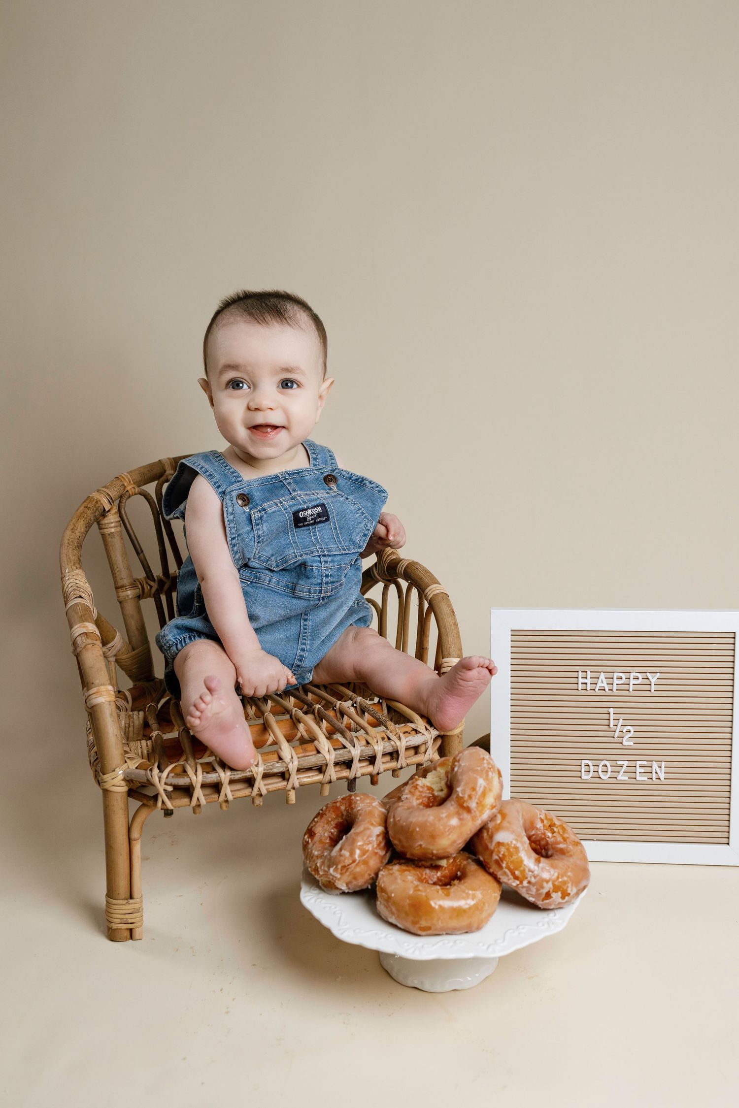 Baby in denim overalls sits on rattan chair next to plate of fresh donuts and letterboard sign.
