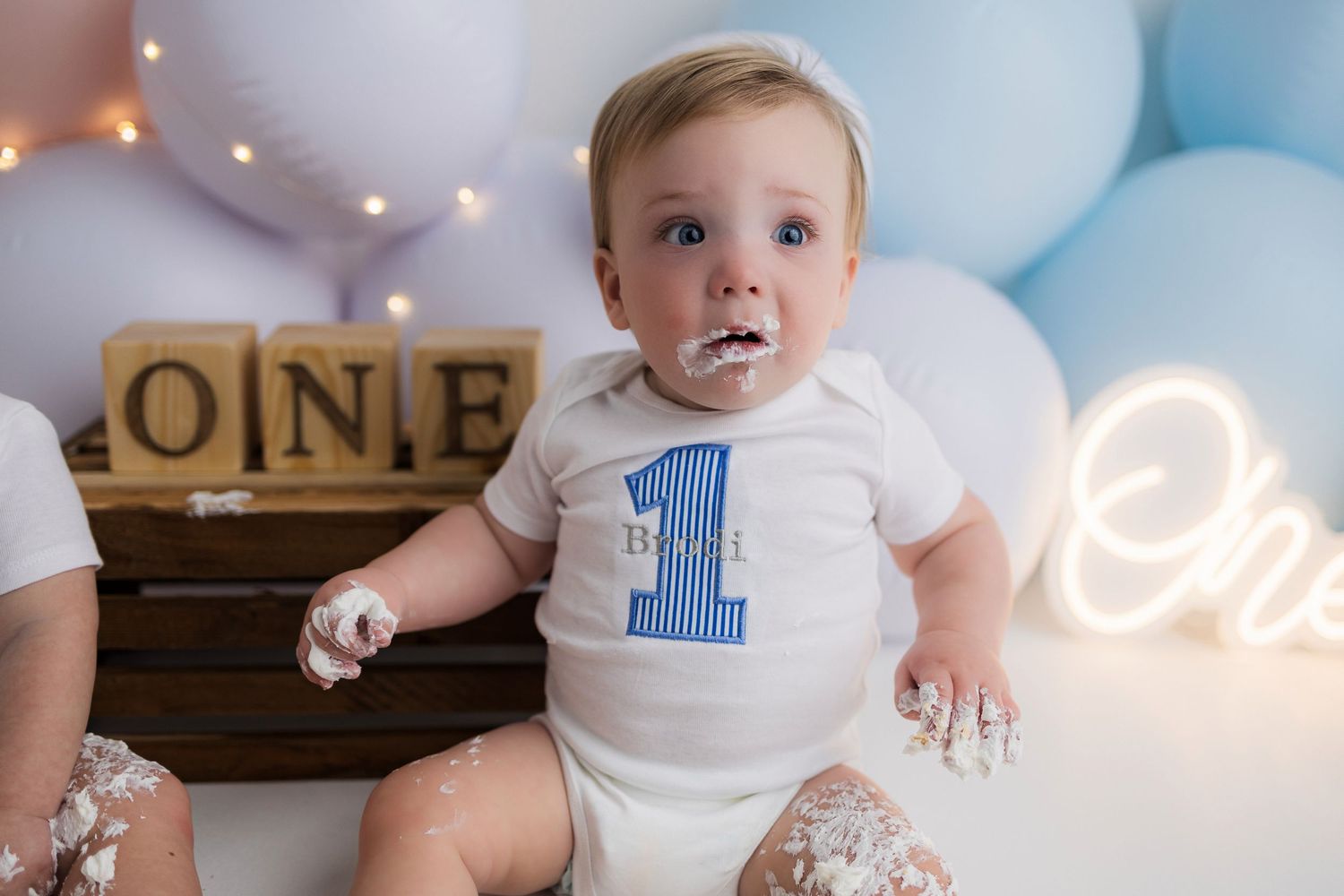 Baby in white first birthday onesie sits amongst blue and white balloons with rustic wooden blocks for cake smash photos.