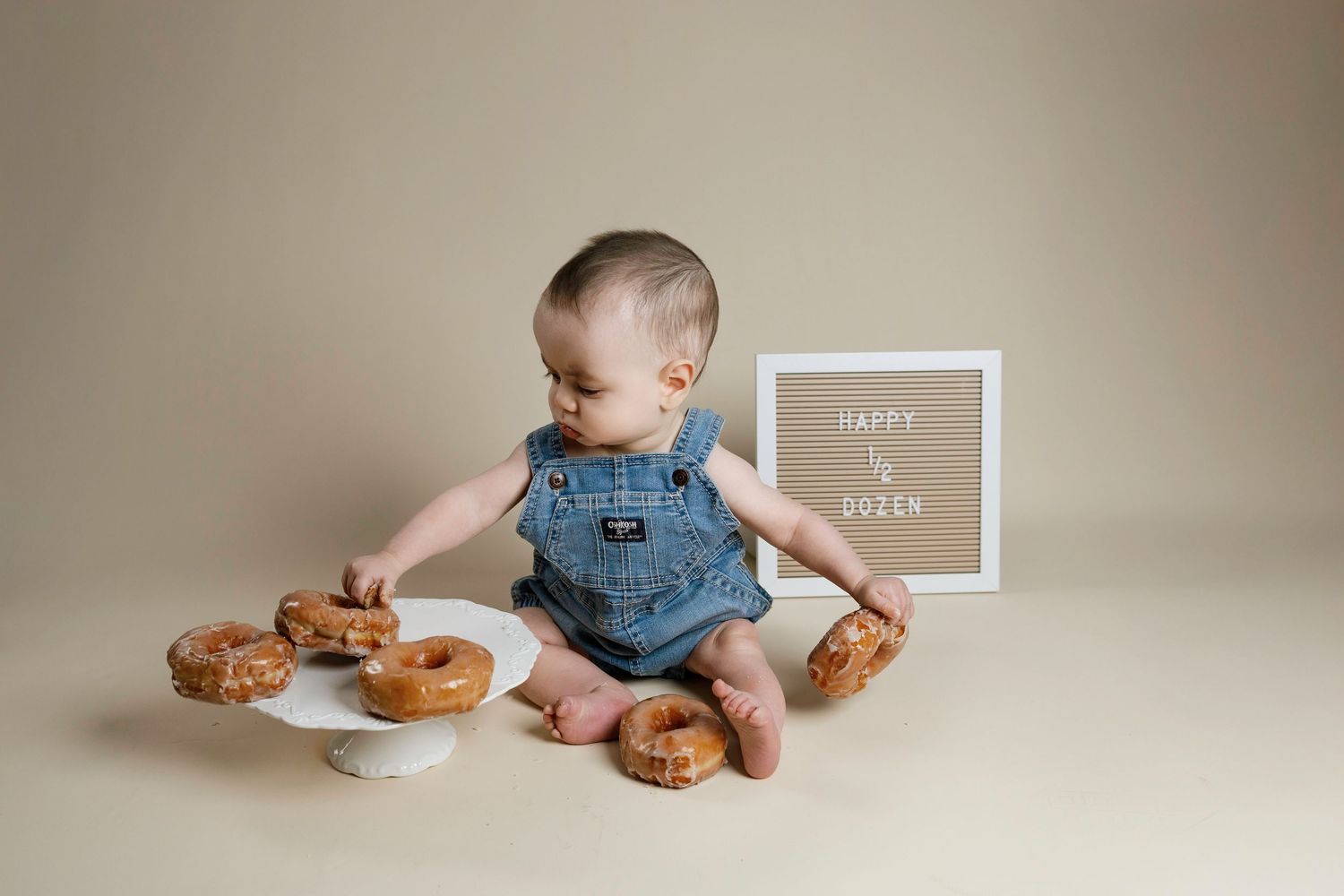 Baby in denim overalls sits on beige backdrop with donuts for first birthday milestone photo session.