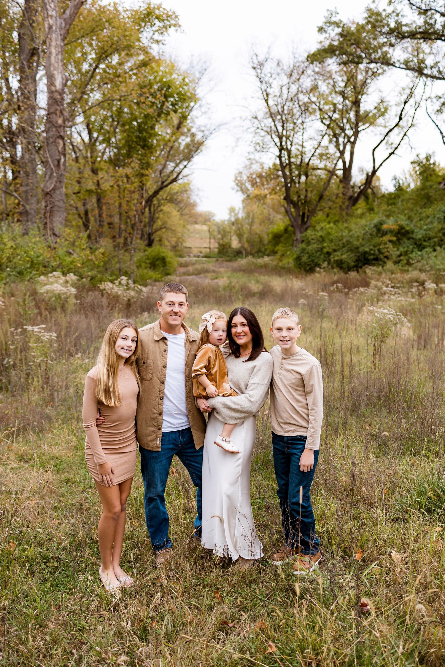 A family poses together in a rustic outdoor setting with tall grass and autumn trees in the background.