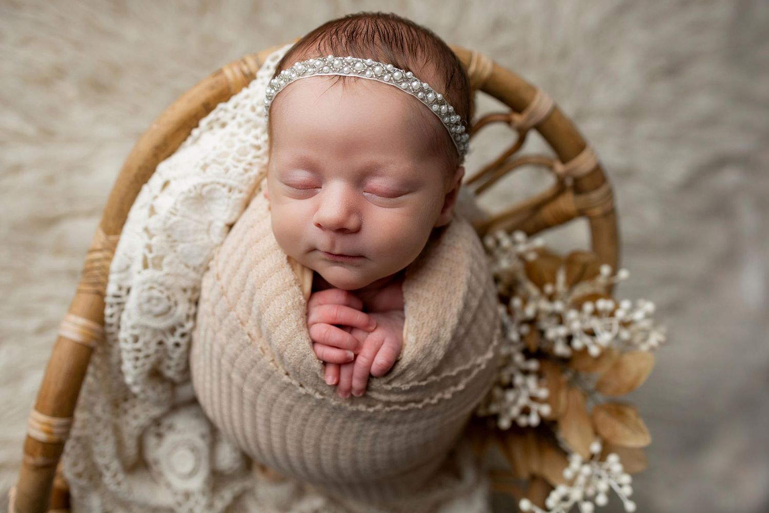 Sleeping newborn baby swaddled in beige knit wrap with delicate white flowers and rustic wooden accents.