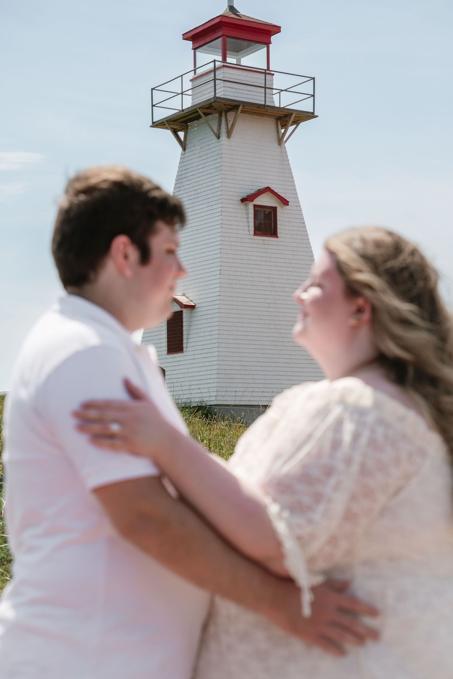 Couple embraces and gazes at each other with a white and red lighthouse in the background on a sunny day.