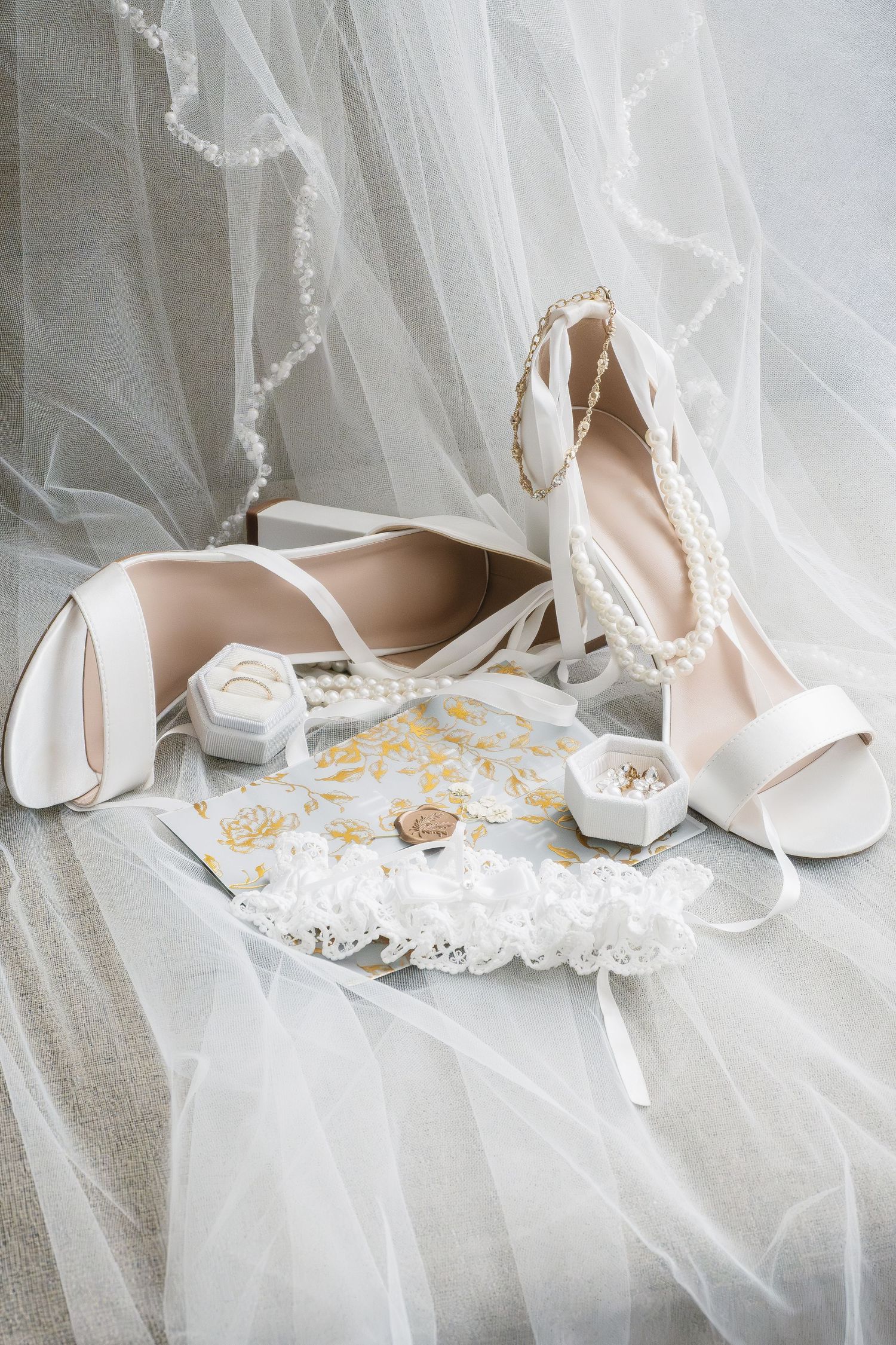 Bridal accessories flat lay with white heels, pearl jewelry, lace garter, and rings on flowing veil.